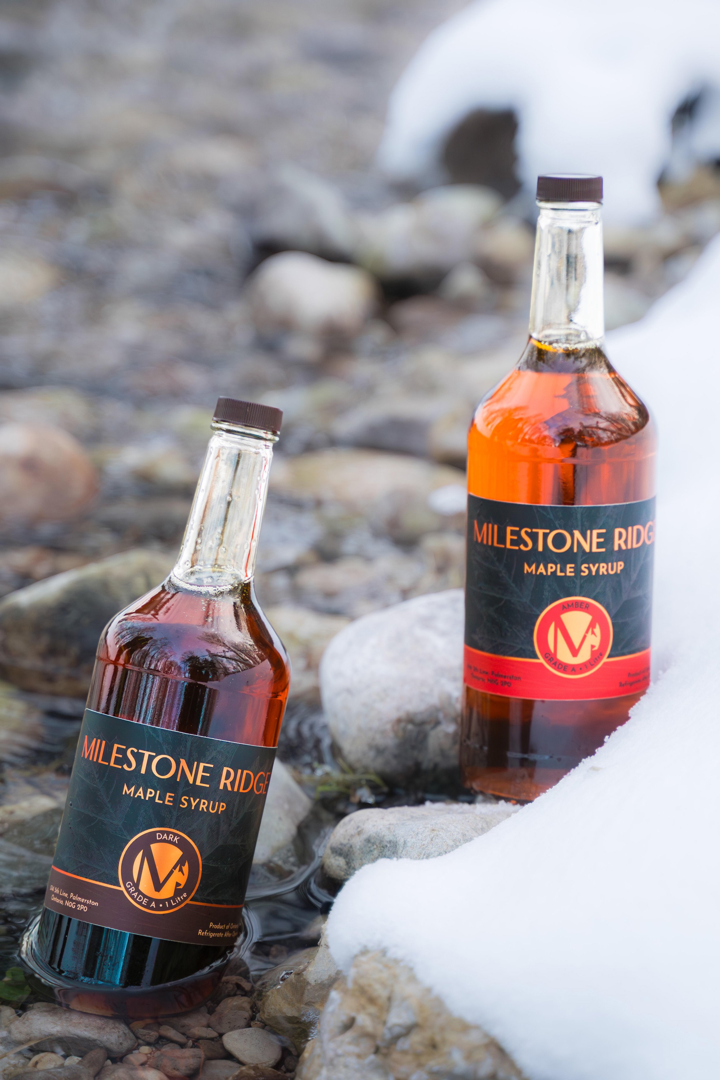 Two glass bottles of Maple Syrup labeled 'Milestone Ridge,' placed on rocky ground with snow in the foreground and blurred rocks and snow in the background. Maple syrup in Palmerston Ontario, buy maple syrup near me, maple syrup Wellington County.