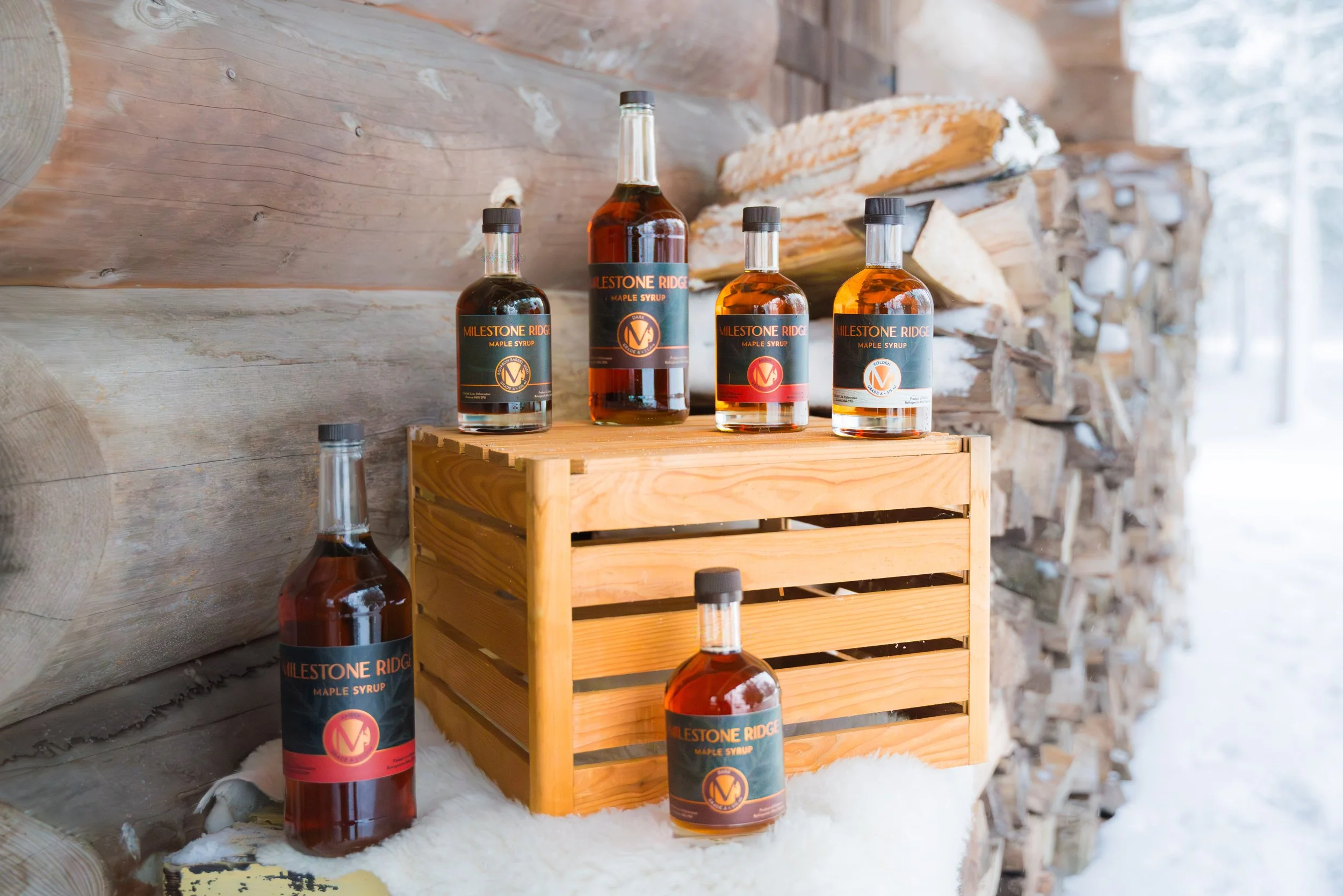 Four bottles of maple syrup labeled "Milestone Ridge" on a wooden surface against a rustic wooden wall. Wholesale maple syrup in Ontario, bulk maple syrup Canada, custom maple syrup gifts, corporate maple syrup favours, maple syrup gift ideas Ontario