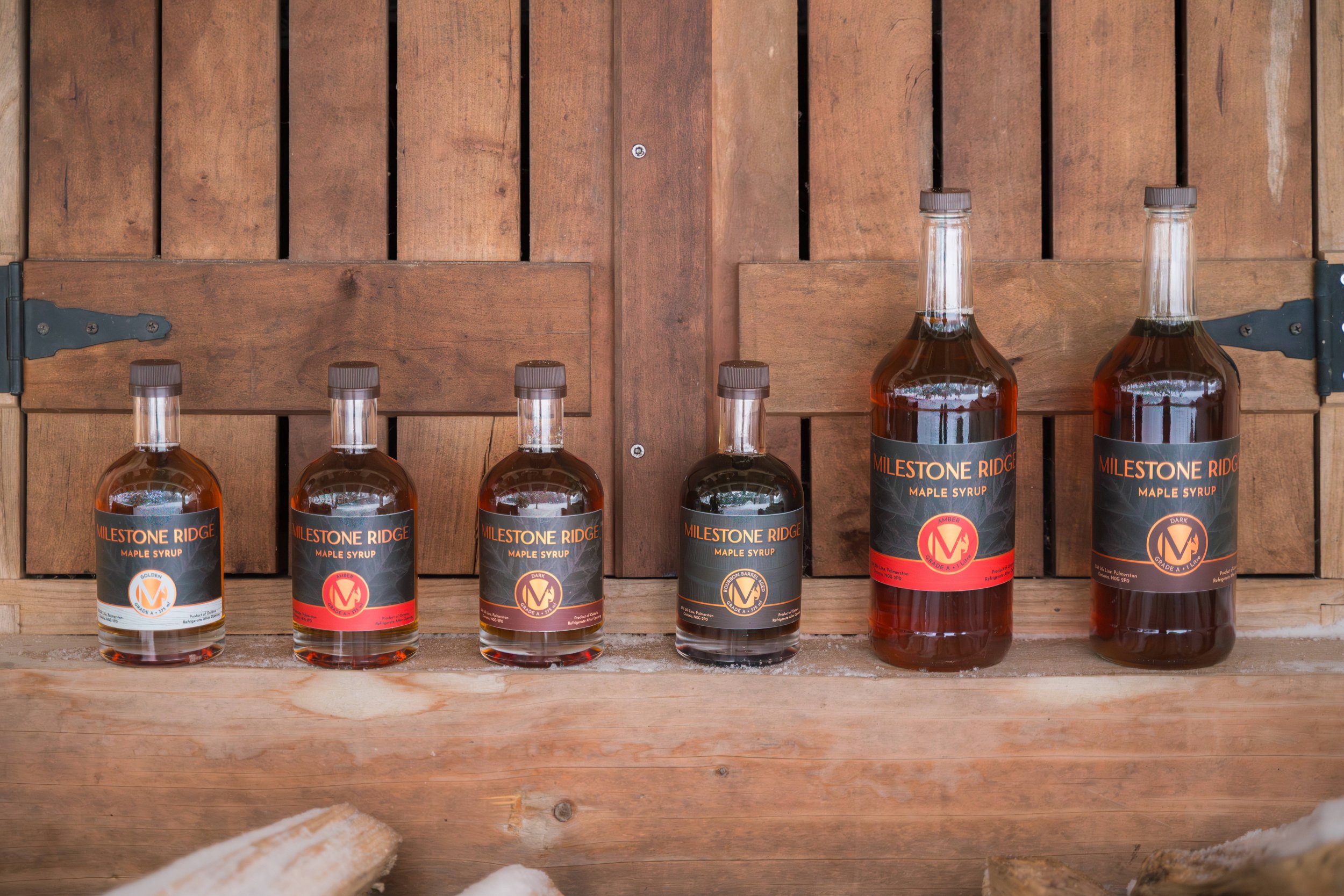 Several bottles of maple syrup labeled "Milestone Ridge" are arranged on a wooden shelf against a wooden backdrop. Maple syrup in Palmerston Ontario, buy maple syrup near me, maple syrup Wellington County, maple syrup Perth County.