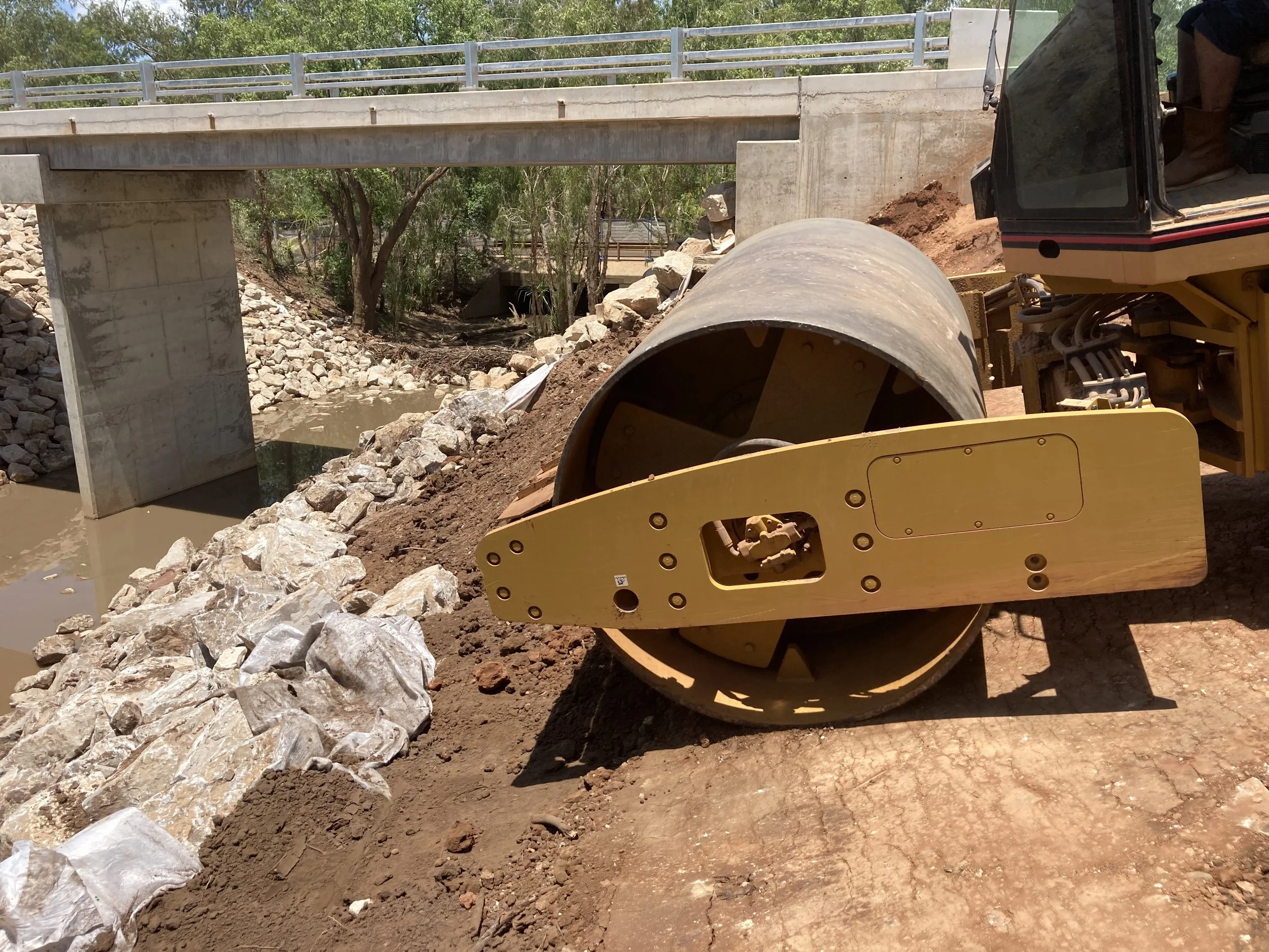 Construction site showing a tunnel boring machine (TBM) next to a small bridge over a creek with trees in the background.