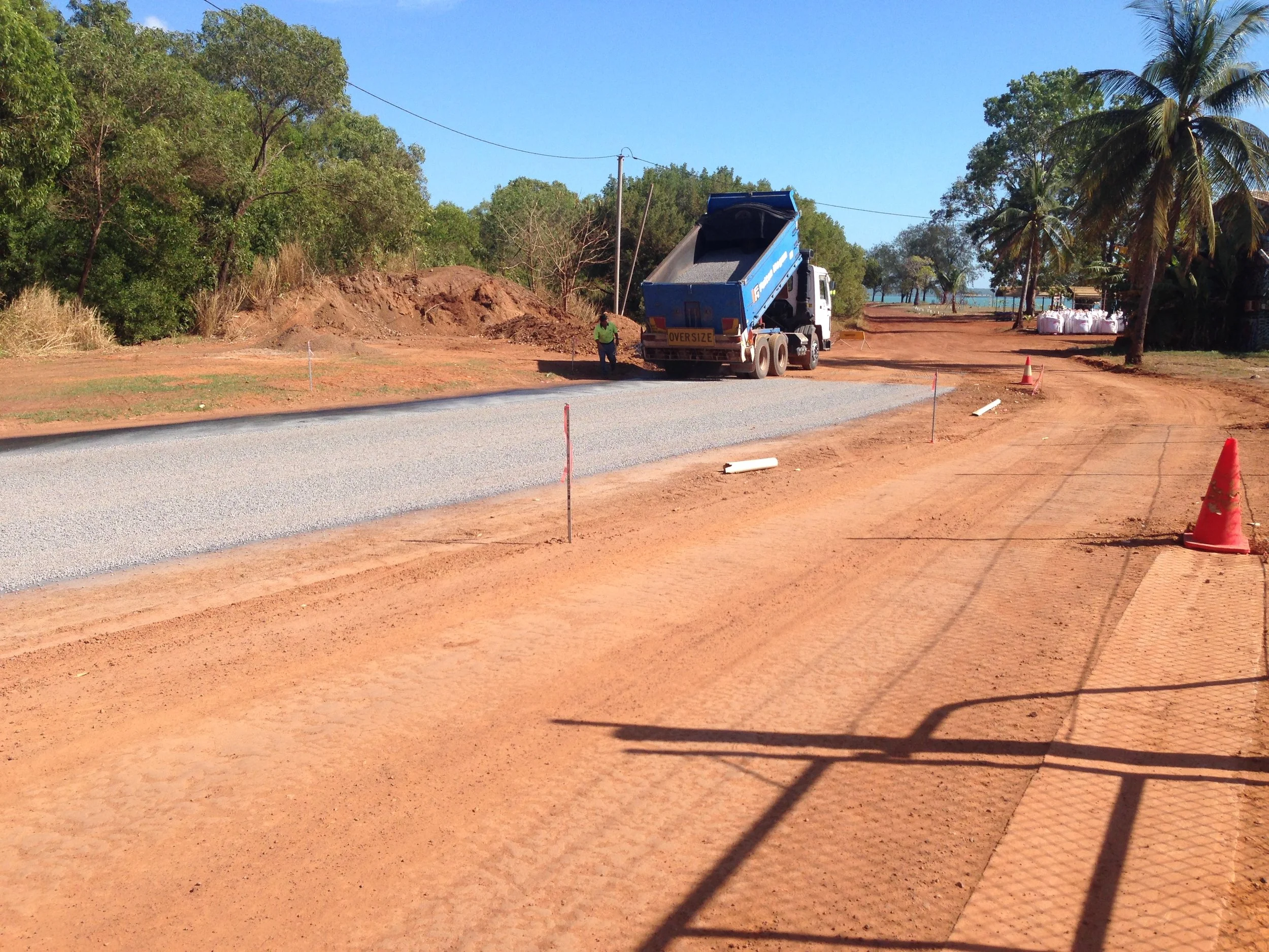 Construction site where a road is being paved with gravel. A dump truck is depositing gravel onto the road, with construction cones and markers along the edges. There are trees and palm trees in the background under a clear blue sky.