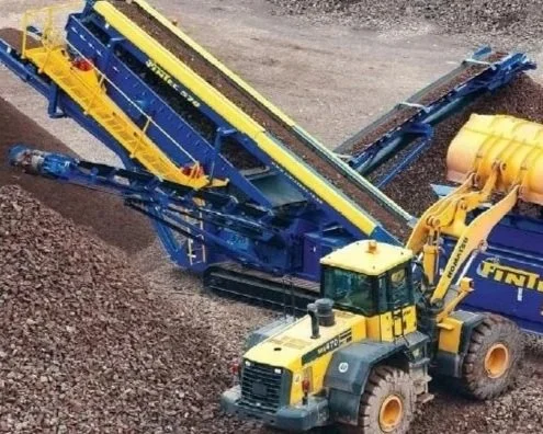 A large yellow and blue industrial crusher or screener machine with a conveyor belt positioned diagonally, and a yellow excavator in the background, working on a gravel or dirt surface.