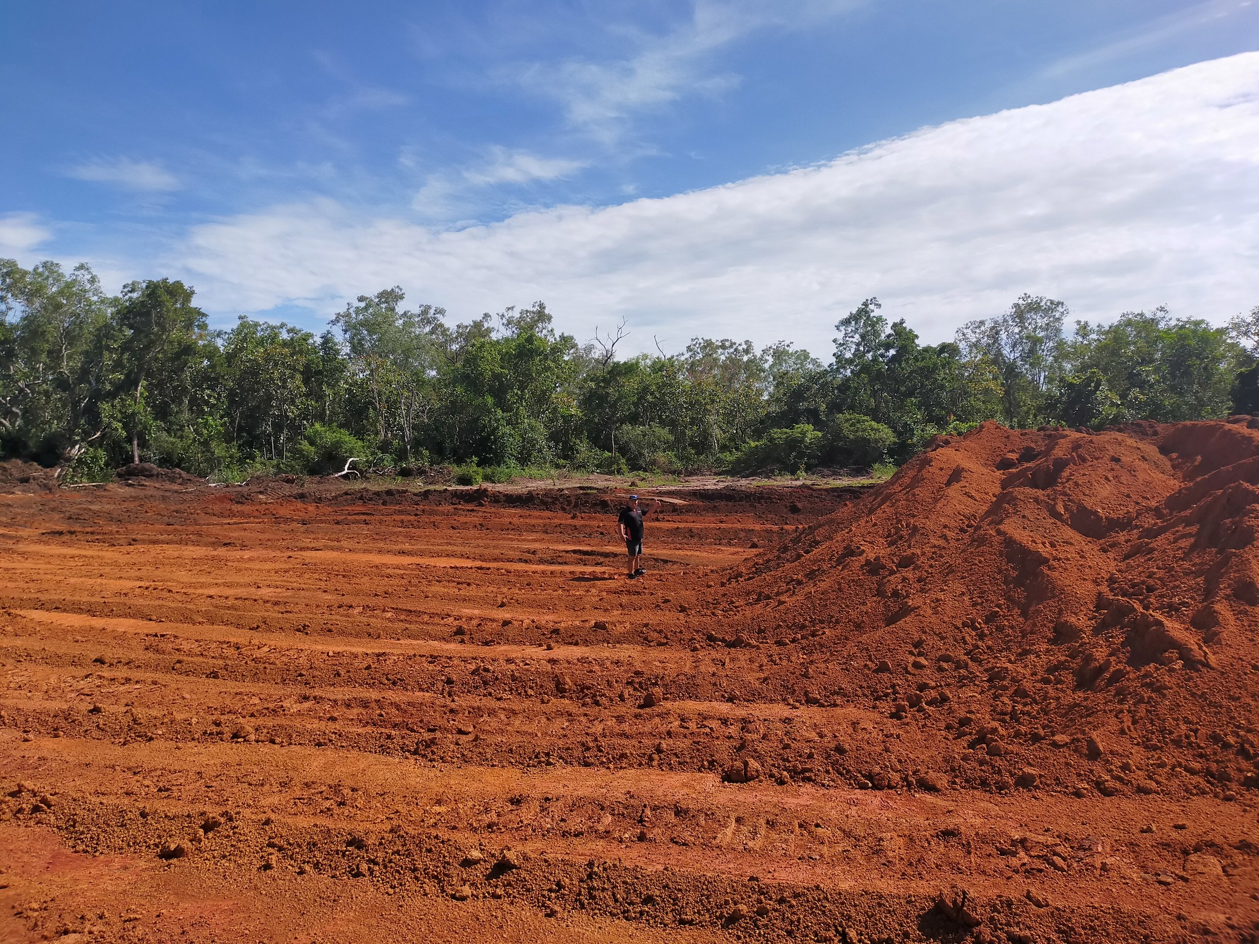 A person standing on red dirt ground with a green forest and blue sky with clouds in the background.