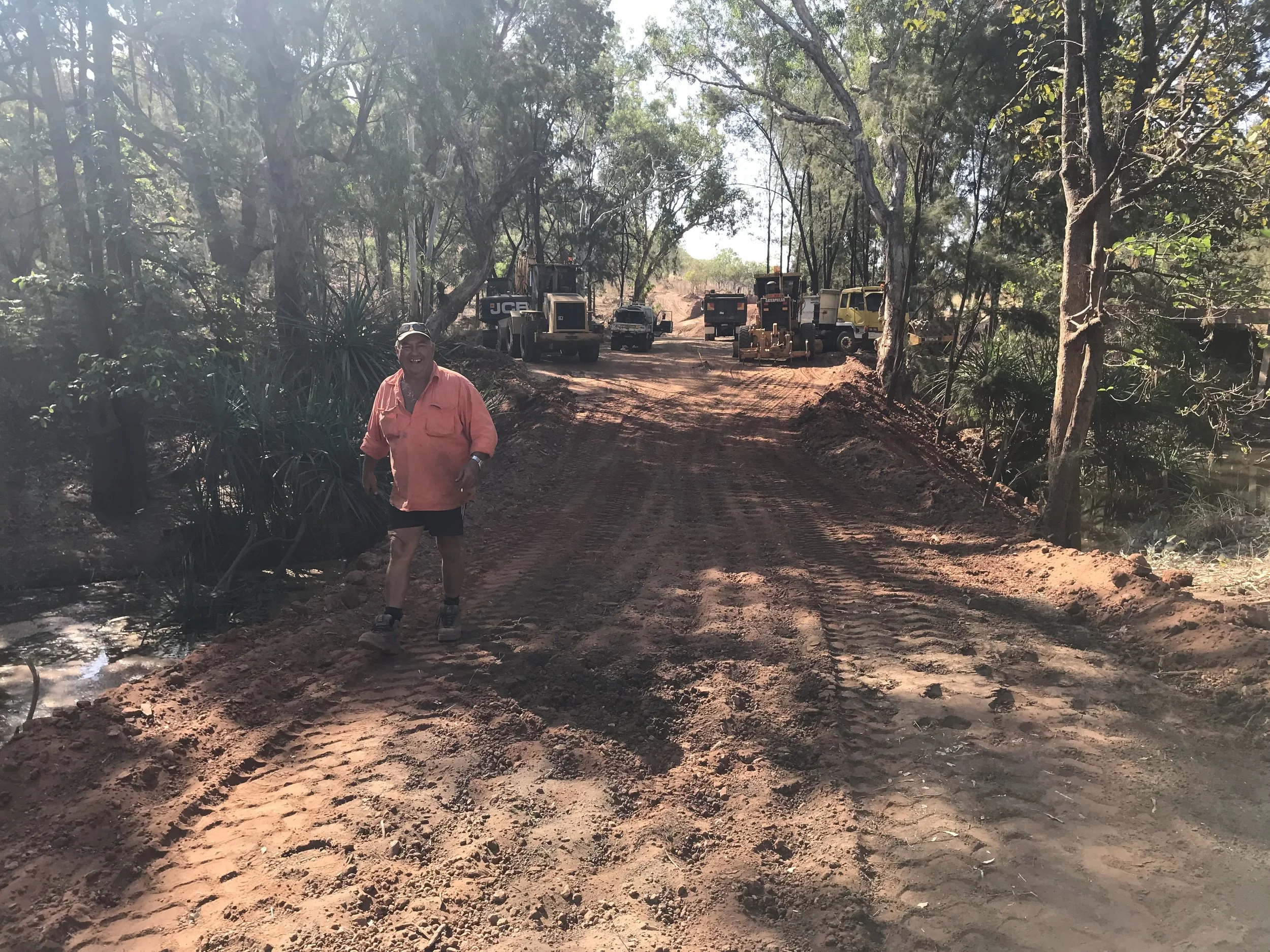 A person in an orange shirt and shorts walking along a dirt path in a wooded area, with construction machinery and trees in the background.
