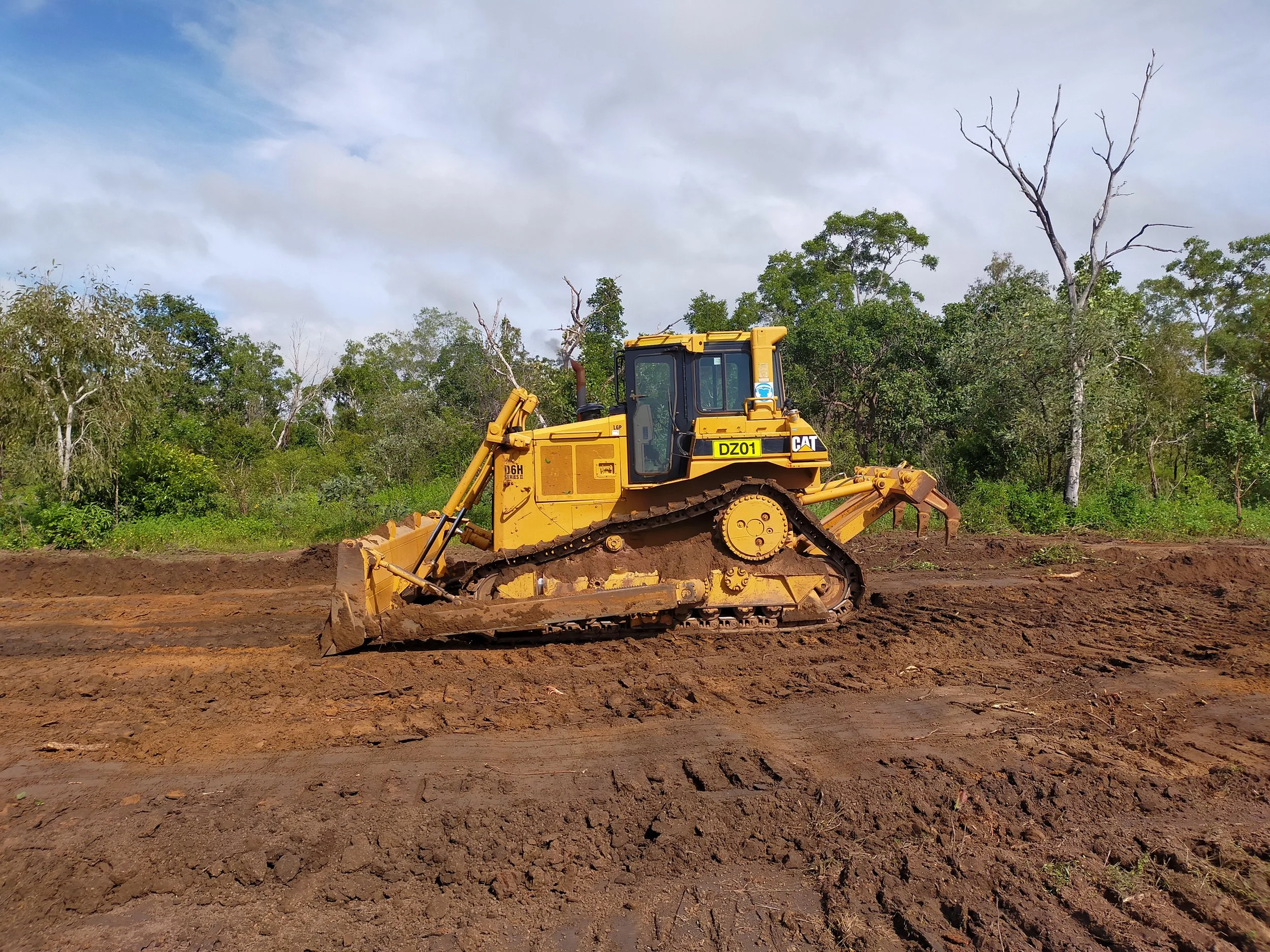 A yellow Caterpillar bulldozer working on a dirt road in a rural area with trees in the background and a partly cloudy sky.