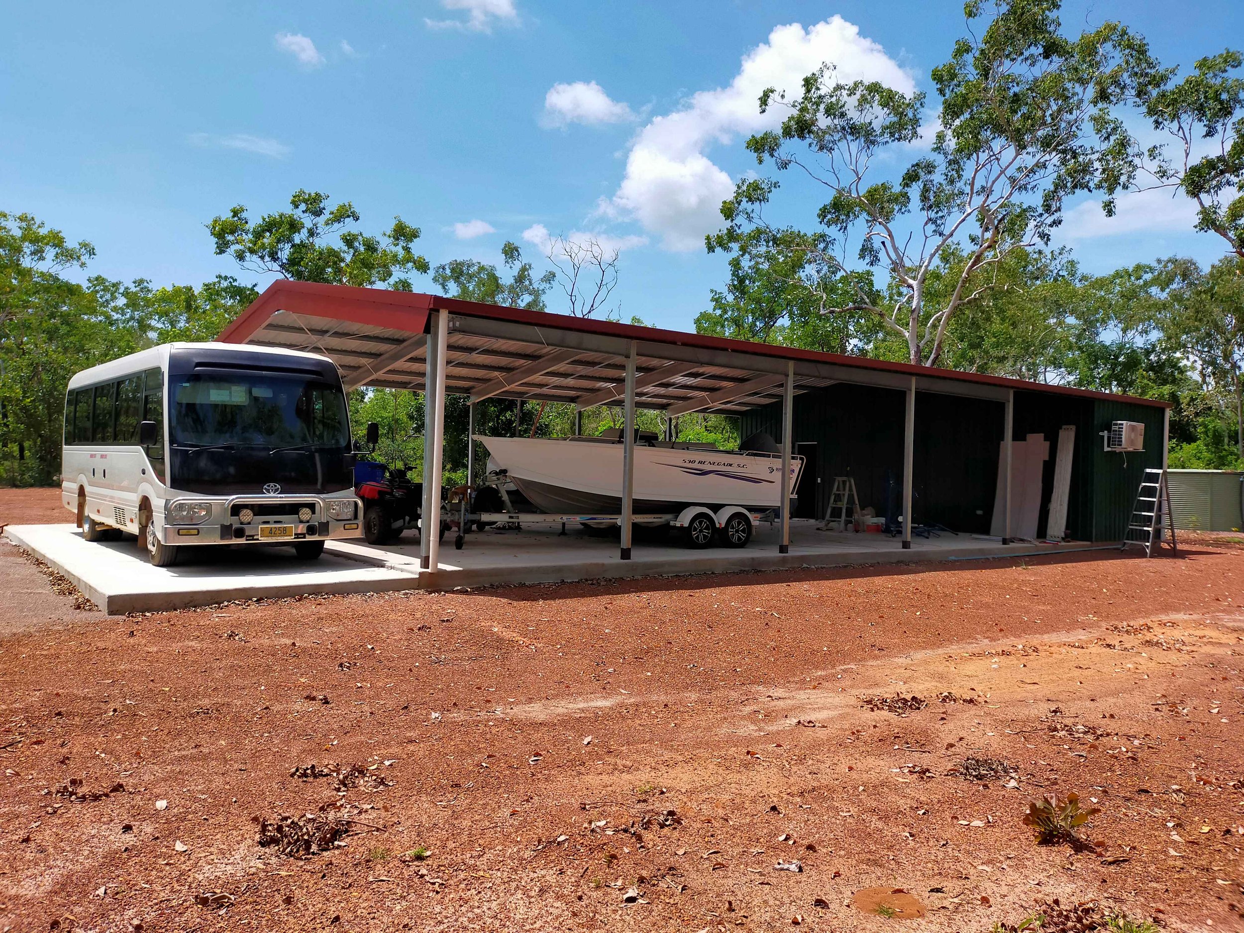 A shed with a red roof, housing a boat on a trailer, a small bus, and a riding mower, surrounded by red dirt and trees under a partly cloudy sky.