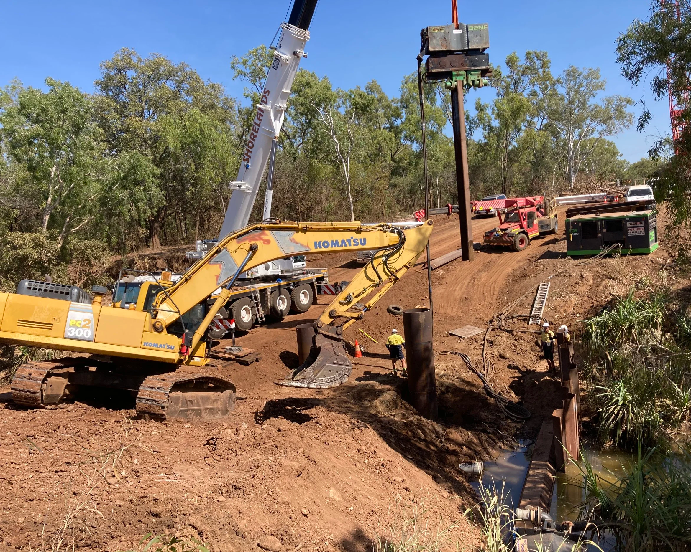 Construction site with a Komatsu excavator, crane, utility trucks, workers, and a power pole being installed near a waterway in a wooded area.