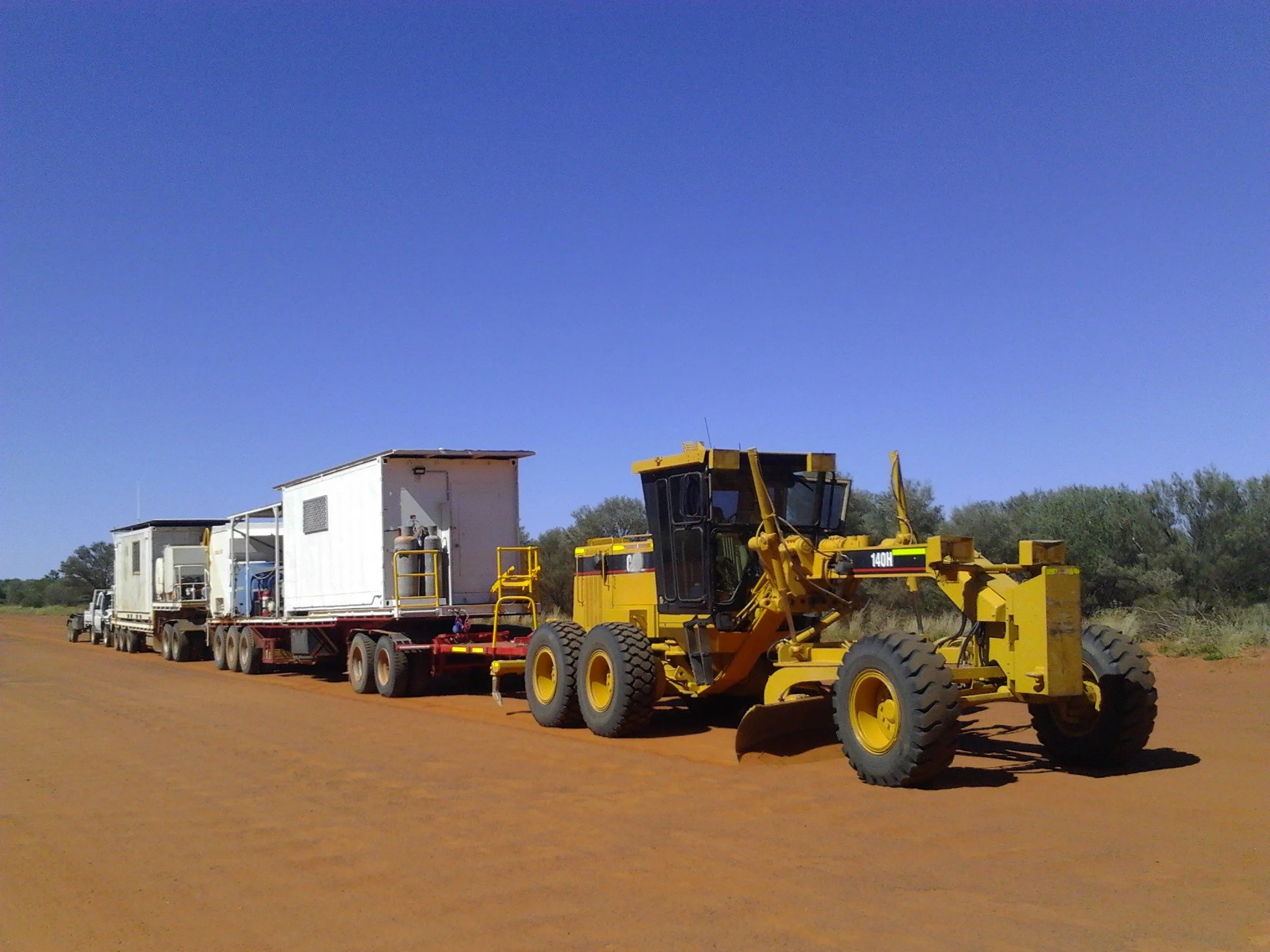 A yellow road grader pulling a flatbed truck with two white containers in a desert landscape with trees in the background and a clear blue sky.