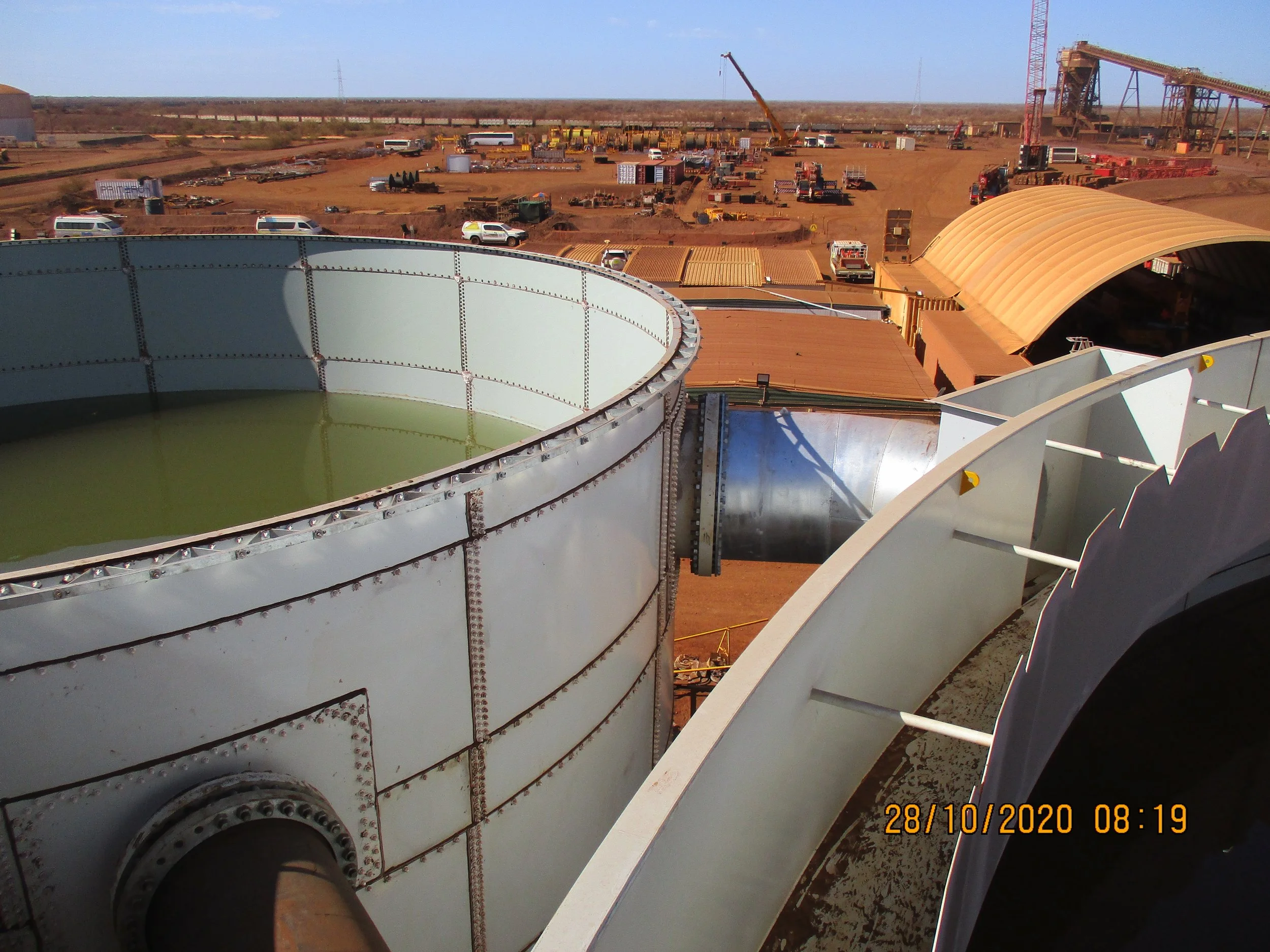 Industrial site with large storage tanks, construction equipment, and vehicles on reddish dirt ground under a clear sky, with a distant power line.