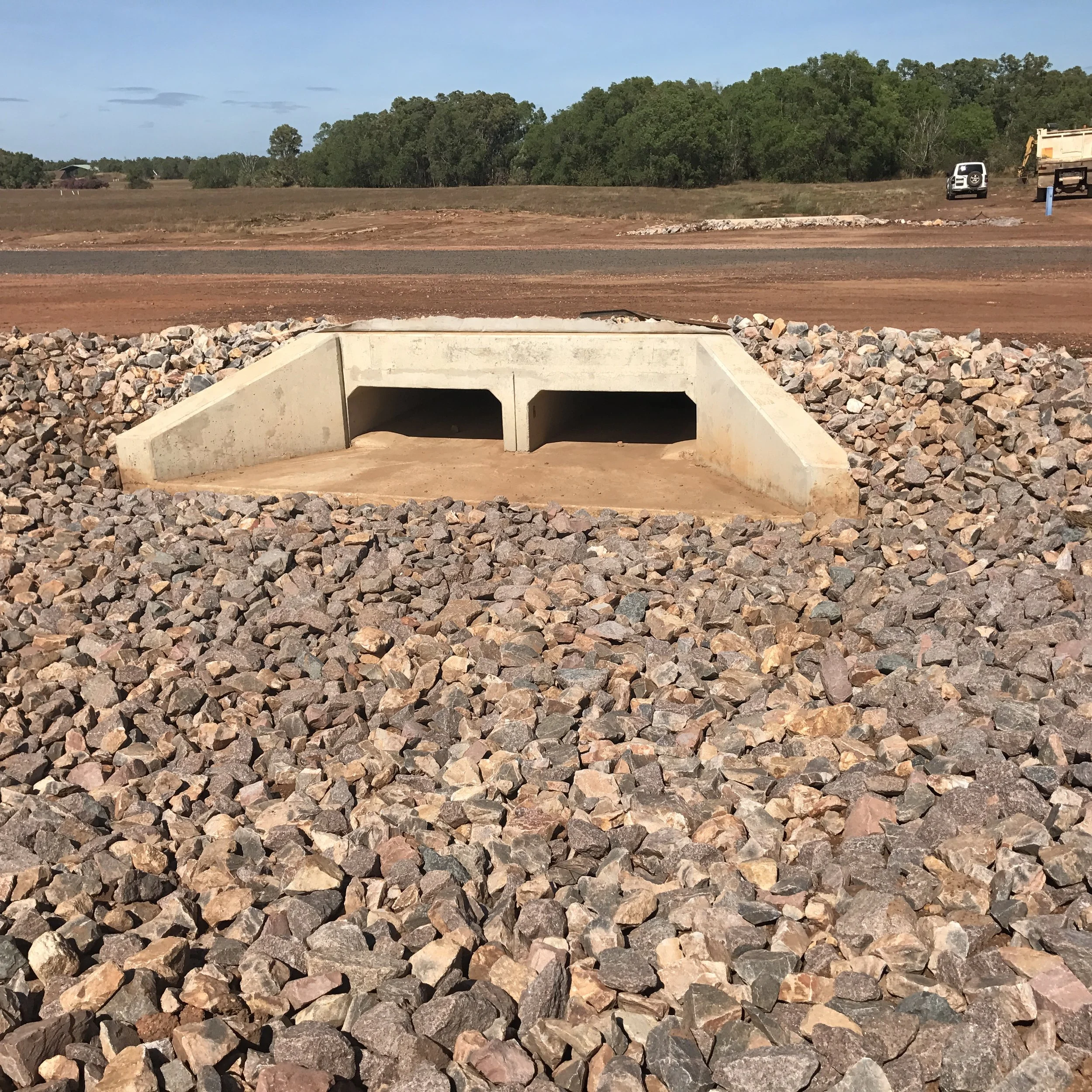 Underground utility access vault with two openings, surrounded by rocks on a construction site, with trees and vehicles in the background.
