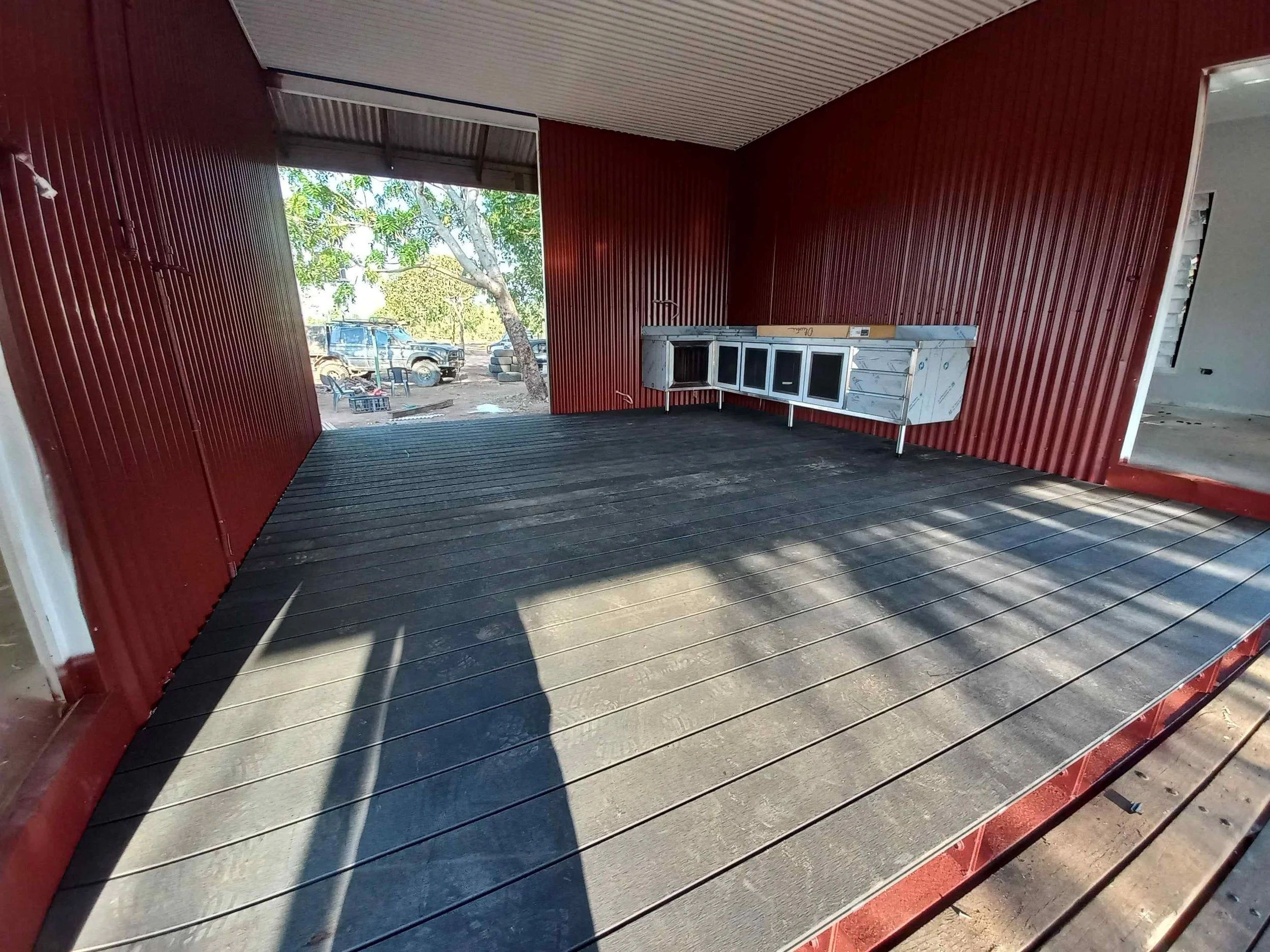 Interior of a room with red corrugated metal walls and a wooden floor, showing an open garage door to the outside with cars and trees visible, and a kitchen counter attached to the wall.