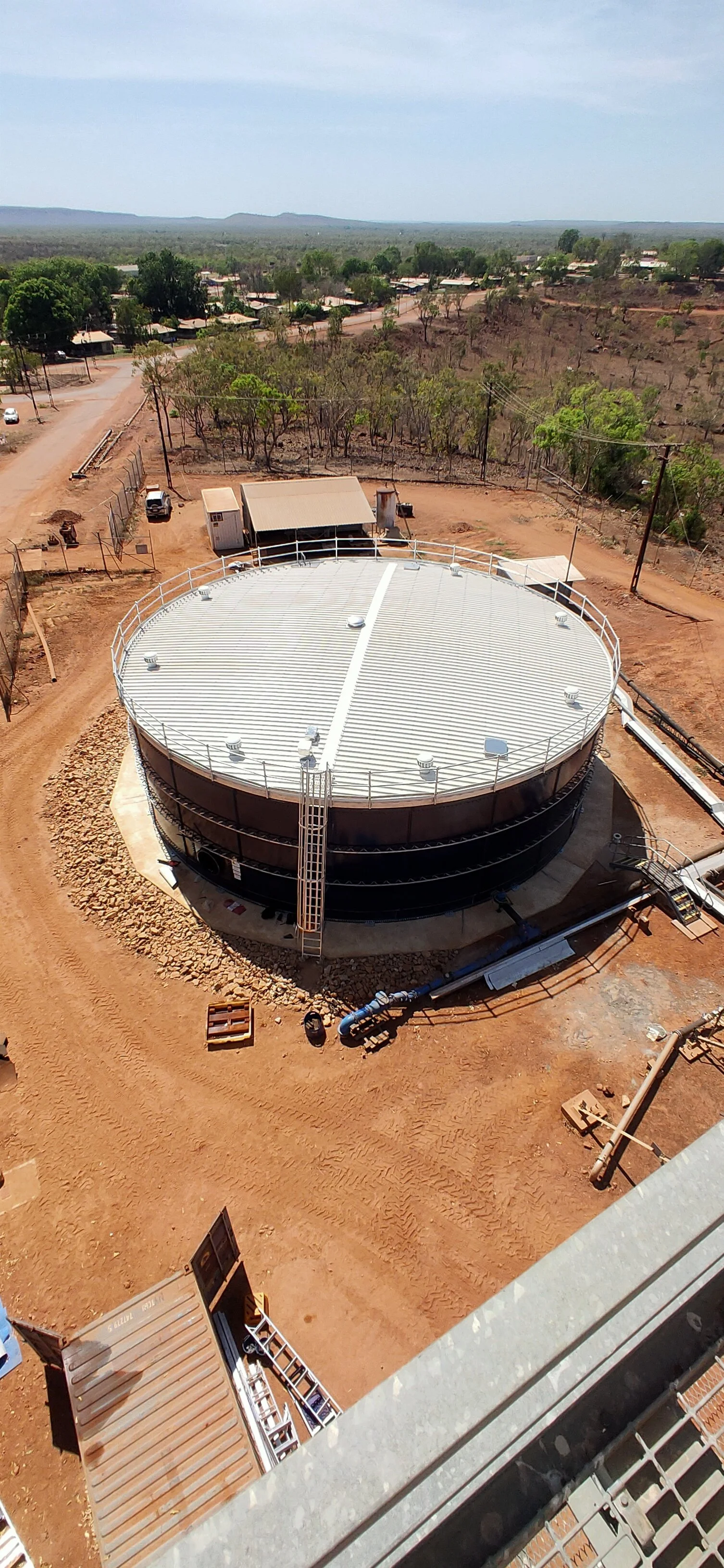 A large, round industrial water tank situated outdoors on reddish dirt ground, surrounded by a fence, with a small building and pipes nearby. In the background, there is an open rural landscape with trees, distant houses, and a light blue sky.