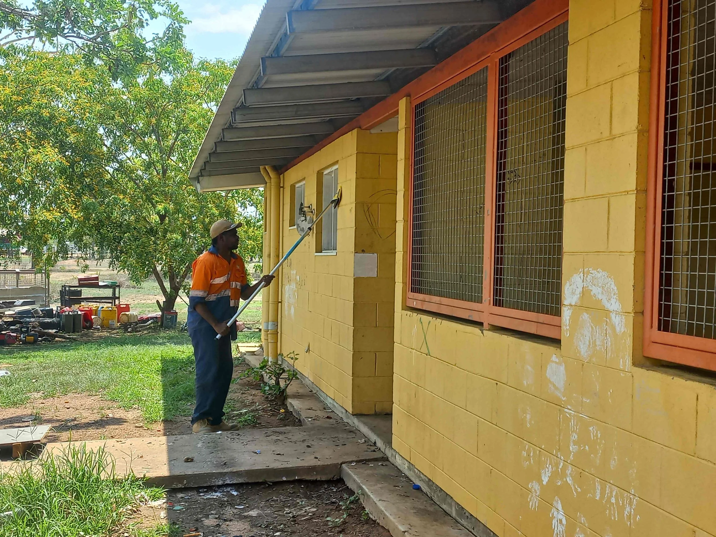 A man in an orange and blue uniform is cleaning the exterior wall of a yellow building with a long tool, possibly a pressure washer, while standing on a concrete pathway in a sunny outdoor environment with green trees and lawn.