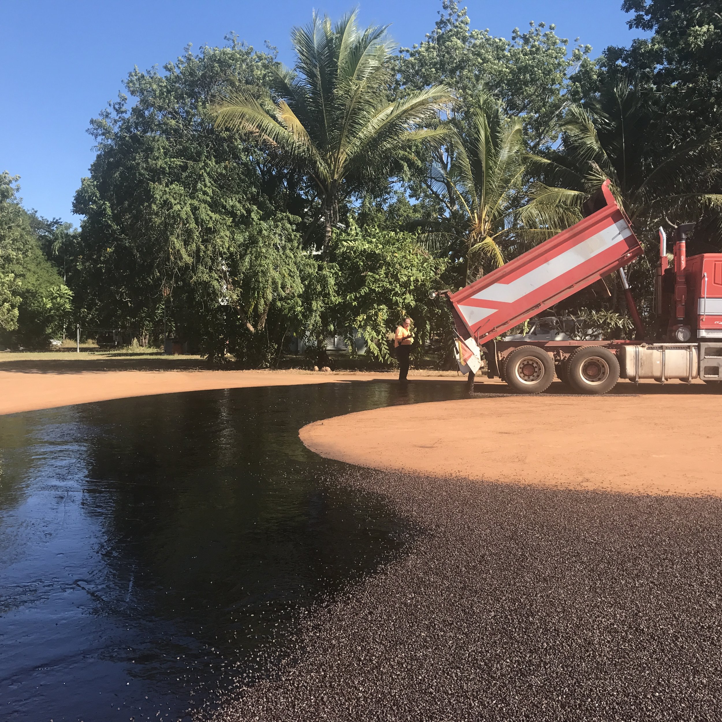 A construction worker standing near a large red truck with its dump bed raised, on a curved area of freshly laid black asphalt, with trees and palm trees in the background under a clear blue sky.