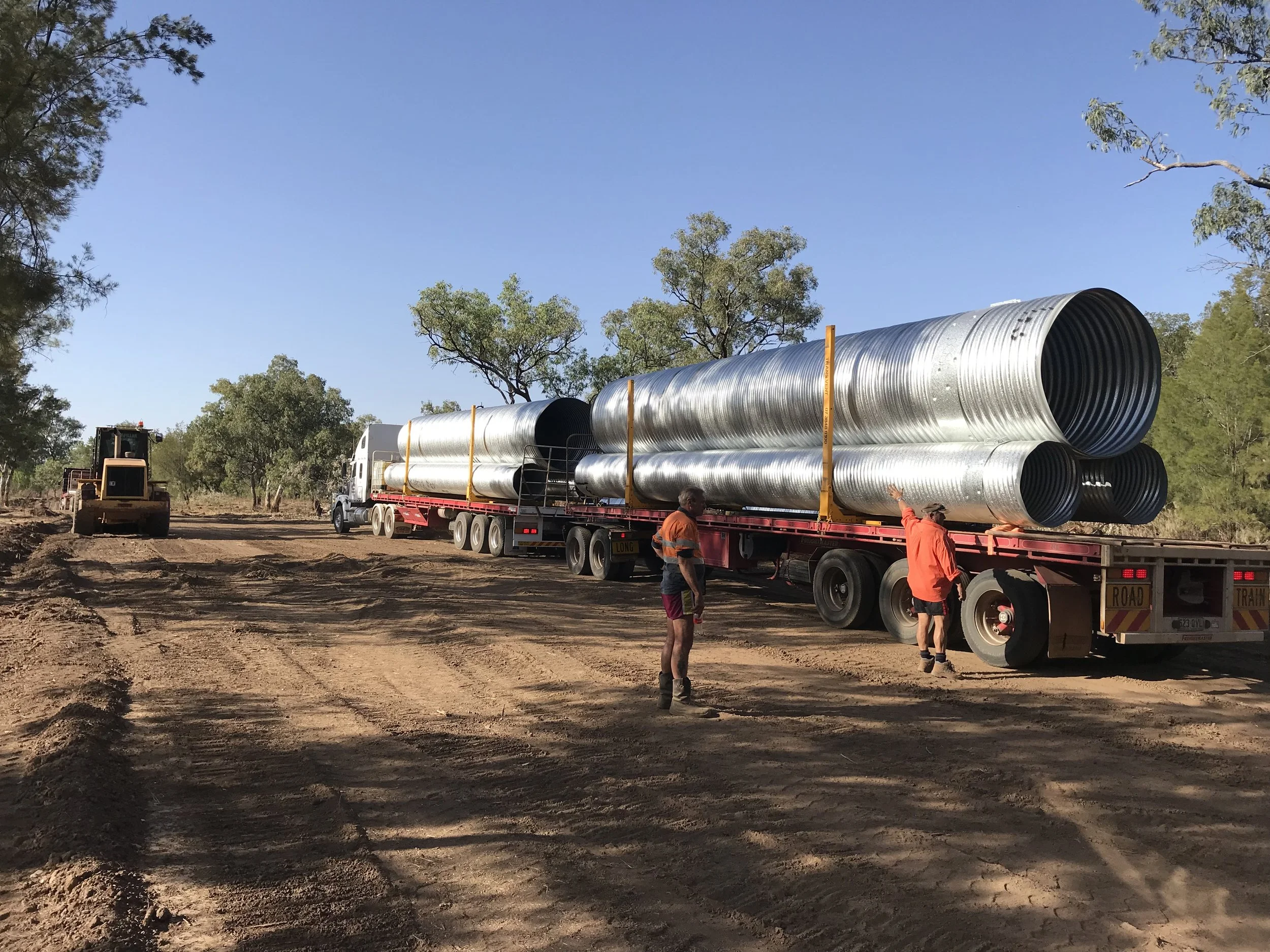 A flatbed truck carrying large metal pipes on a dirt construction site, with two workers in orange safety shirts and a bulldozer in the background, under a clear blue sky and surrounded by trees.