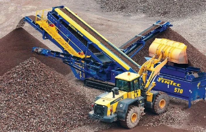 A large yellow bucket wheel excavator loading rocks into a blue conveyor belt system at a quarry site.