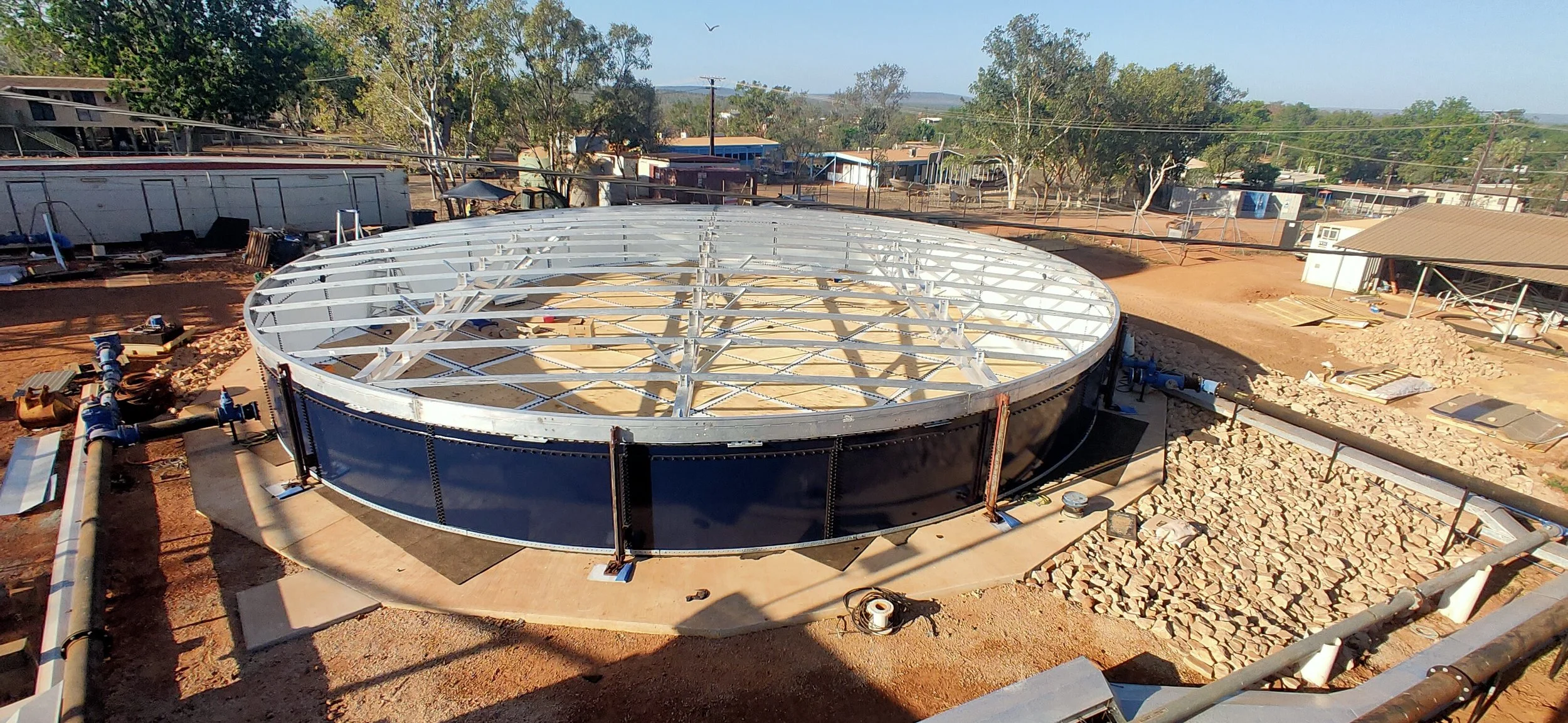 Construction site with a large circular metal structure being assembled, surrounded by construction materials, pipes, and equipment, in a dry area with dirt and sparse trees.