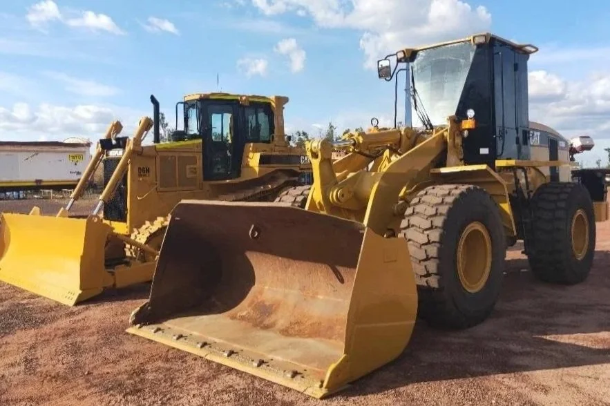 Two large yellow construction bulldozers with wide blades on front, parked on a dirt lot under a partly cloudy sky.