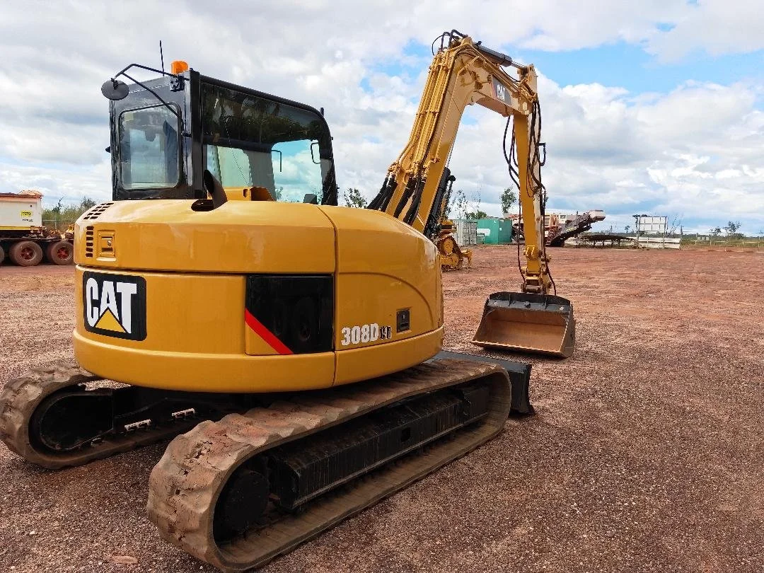 Yellow Caterpillar 308DCR compact excavator with a black cab, sitting on a dirt lot with construction equipment in the background and a partly cloudy sky.