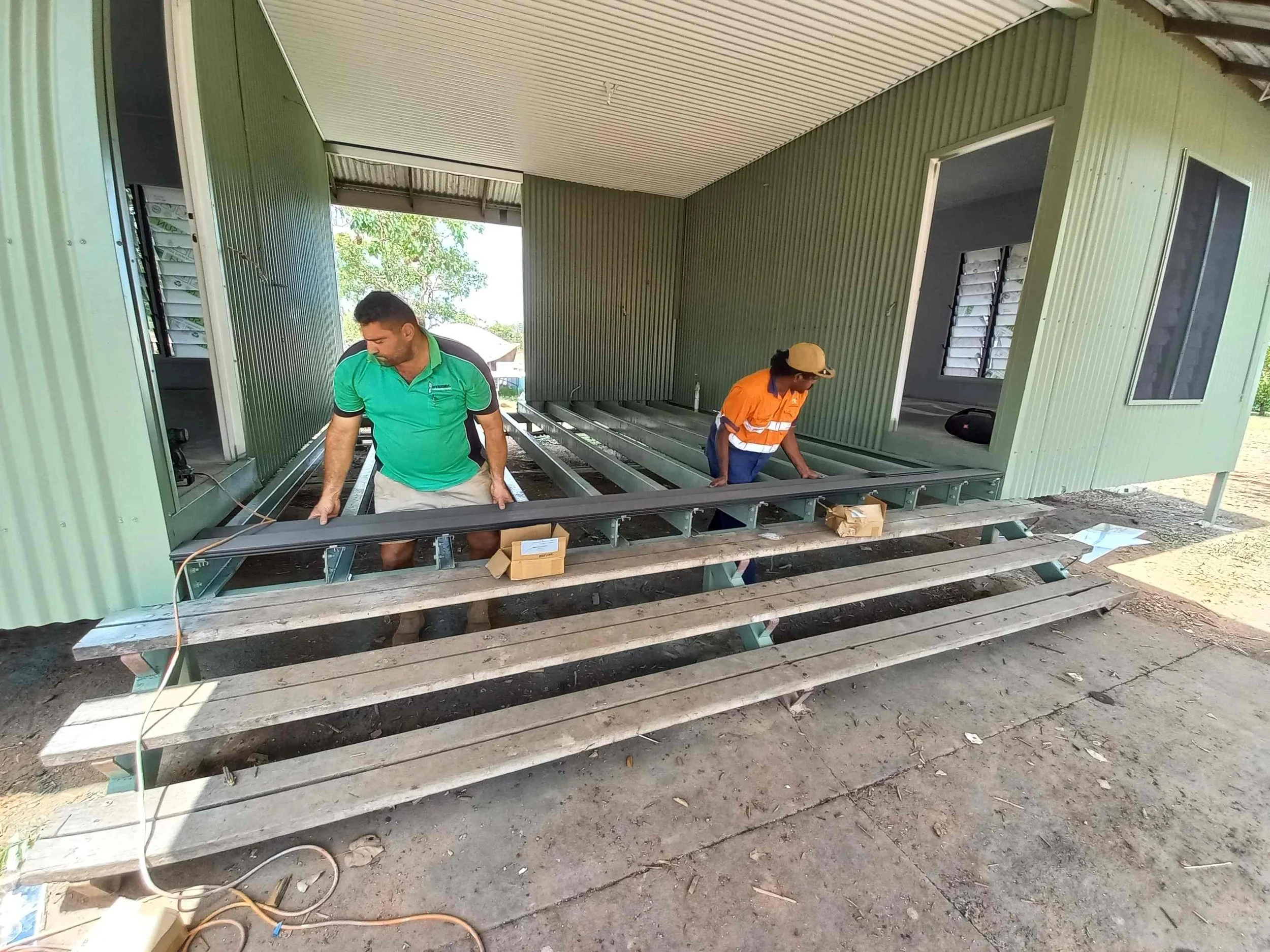 Two workers installing a metal ramp outside a green metal building. One worker wears a green shirt and beige shorts, and the other wears an orange safety vest and a beige hat. They are adjusting the ramp on wooden supports, with construction tools and materials nearby.