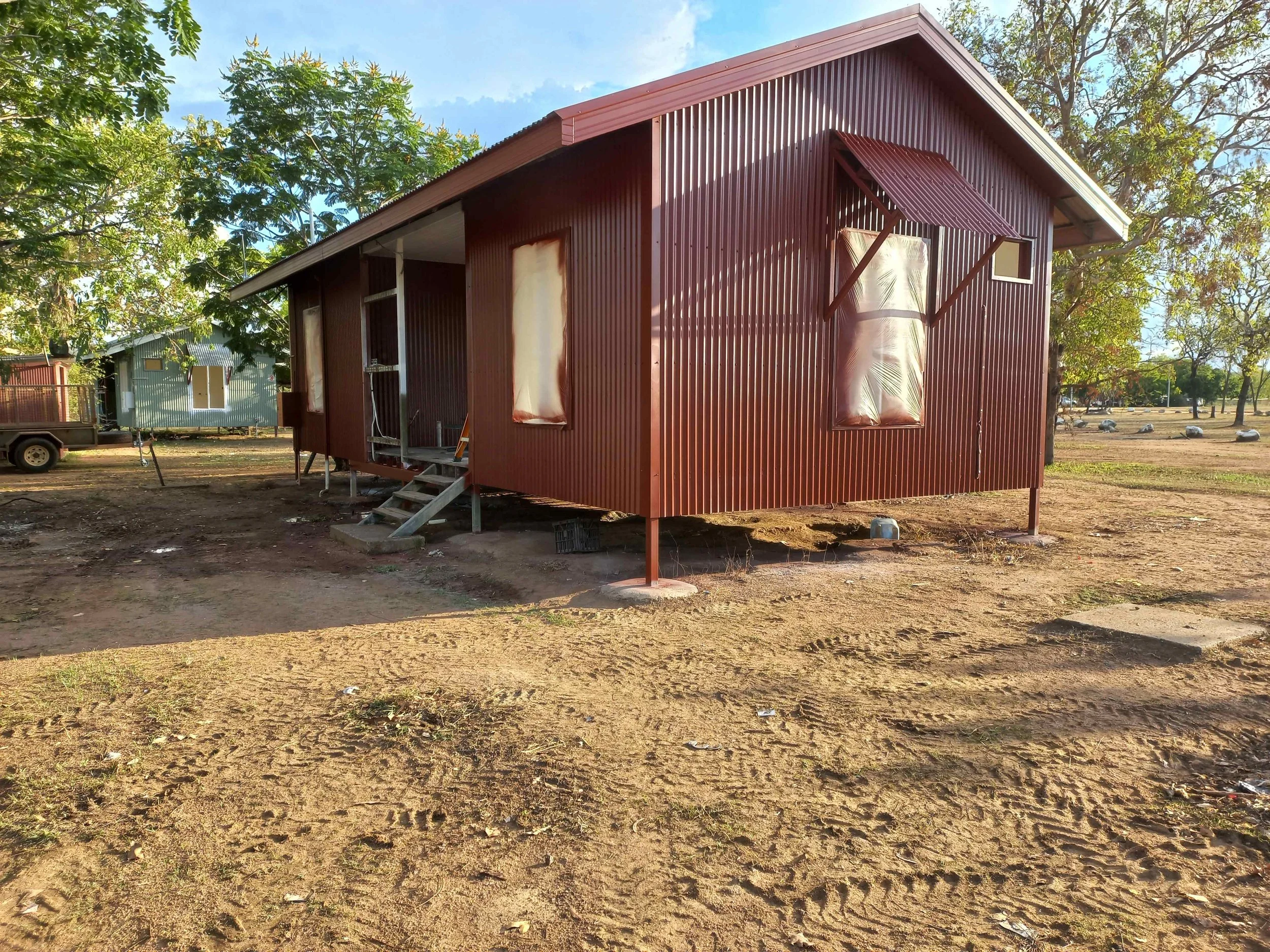 A small, elevated red metal house with a staircase and small awning in an open dirt area with trees in the background.