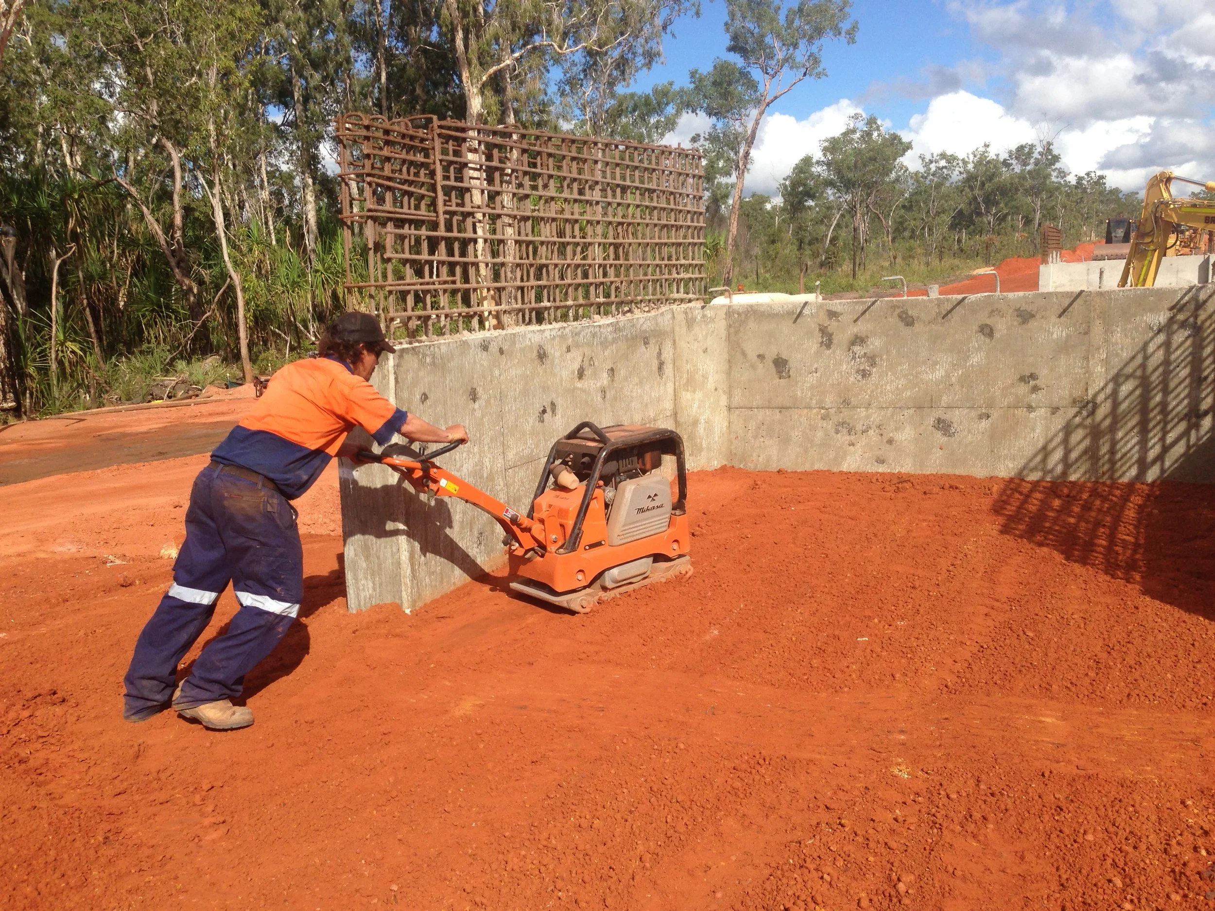 Construction worker operating a compactor on a construction site with red soil, concrete walls, and construction equipment in the background.