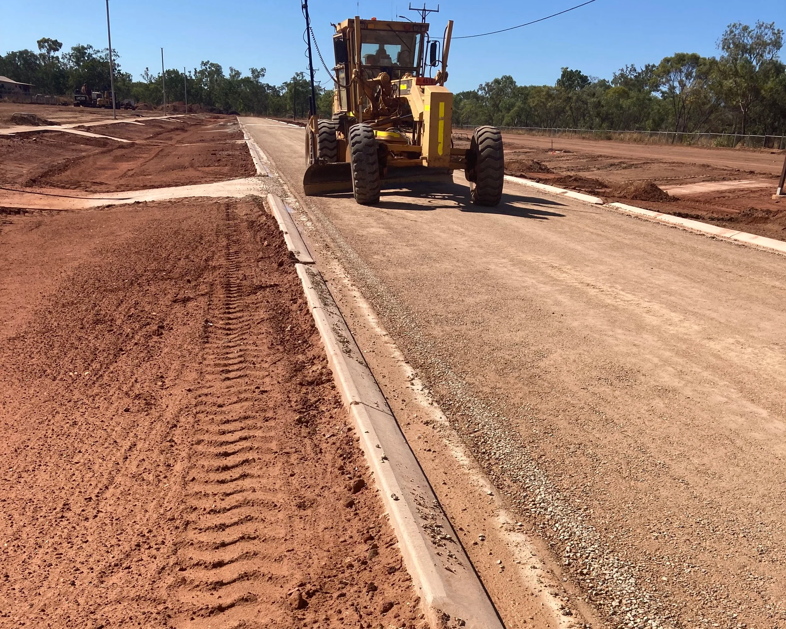 A dirt road under construction with a yellow road grader leveling the surface, surrounded by cleared land and trees in the background.