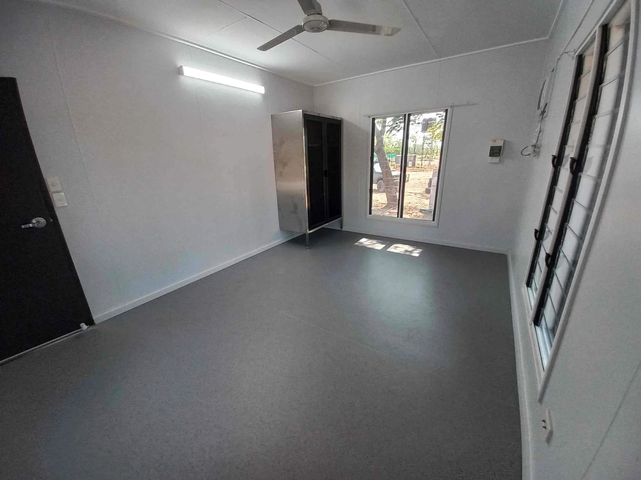 Empty room with white walls, a grey floor, a ceiling fan, a sliding glass door showing outdoor trees and ground, black cabinet, a ceiling-mounted light, a wall-mounted thermostat, and a ladder leaning against the wall.