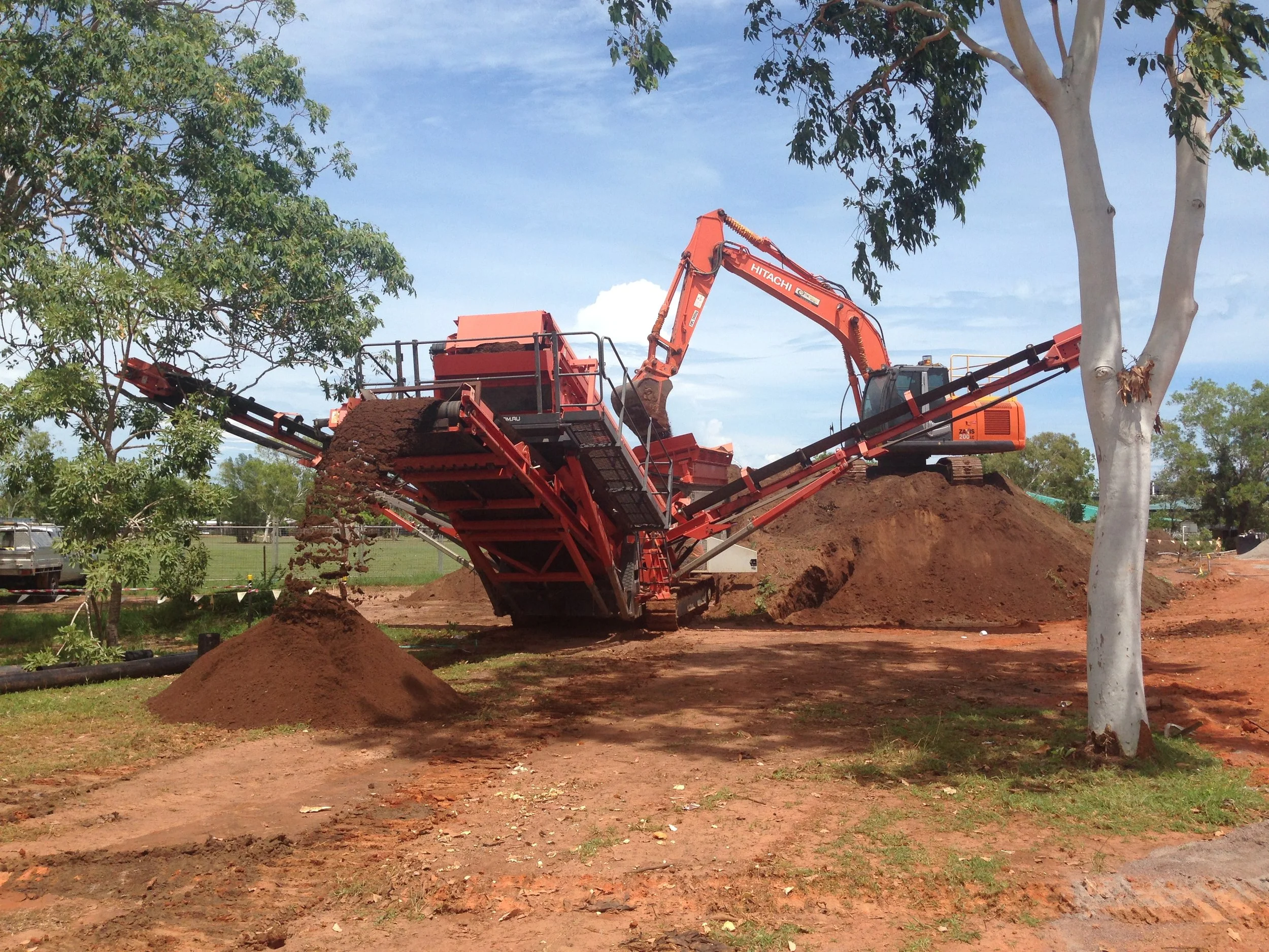 An orange excavator and a large red earth-moving machine are working on a construction site surrounded by trees and parked vehicles. The excavator is lifting dirt, pouring it onto a pile.
