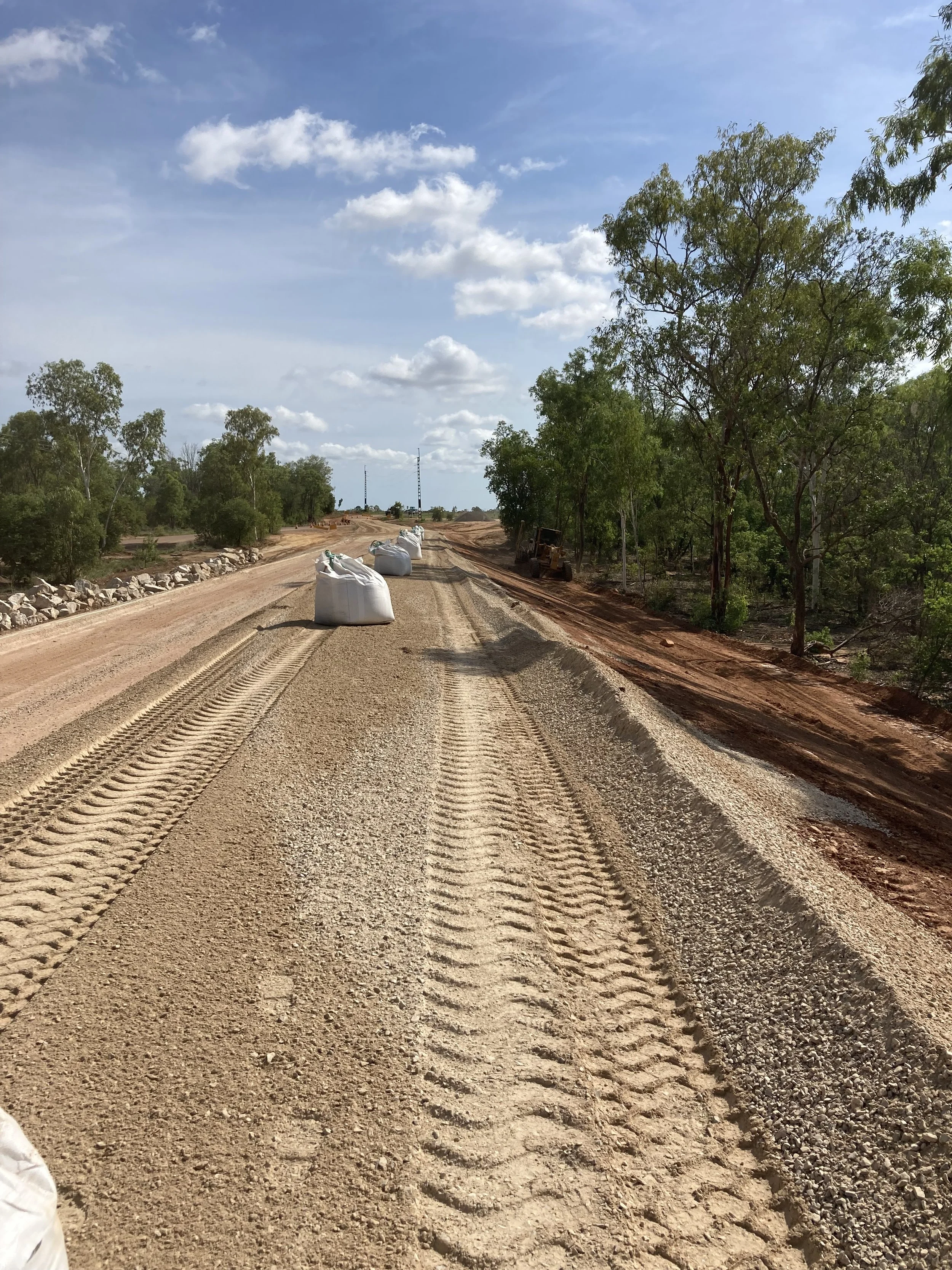 Construction site with a partially paved dirt road, white sandbags, and construction equipment, surrounded by green trees under a partly cloudy sky.