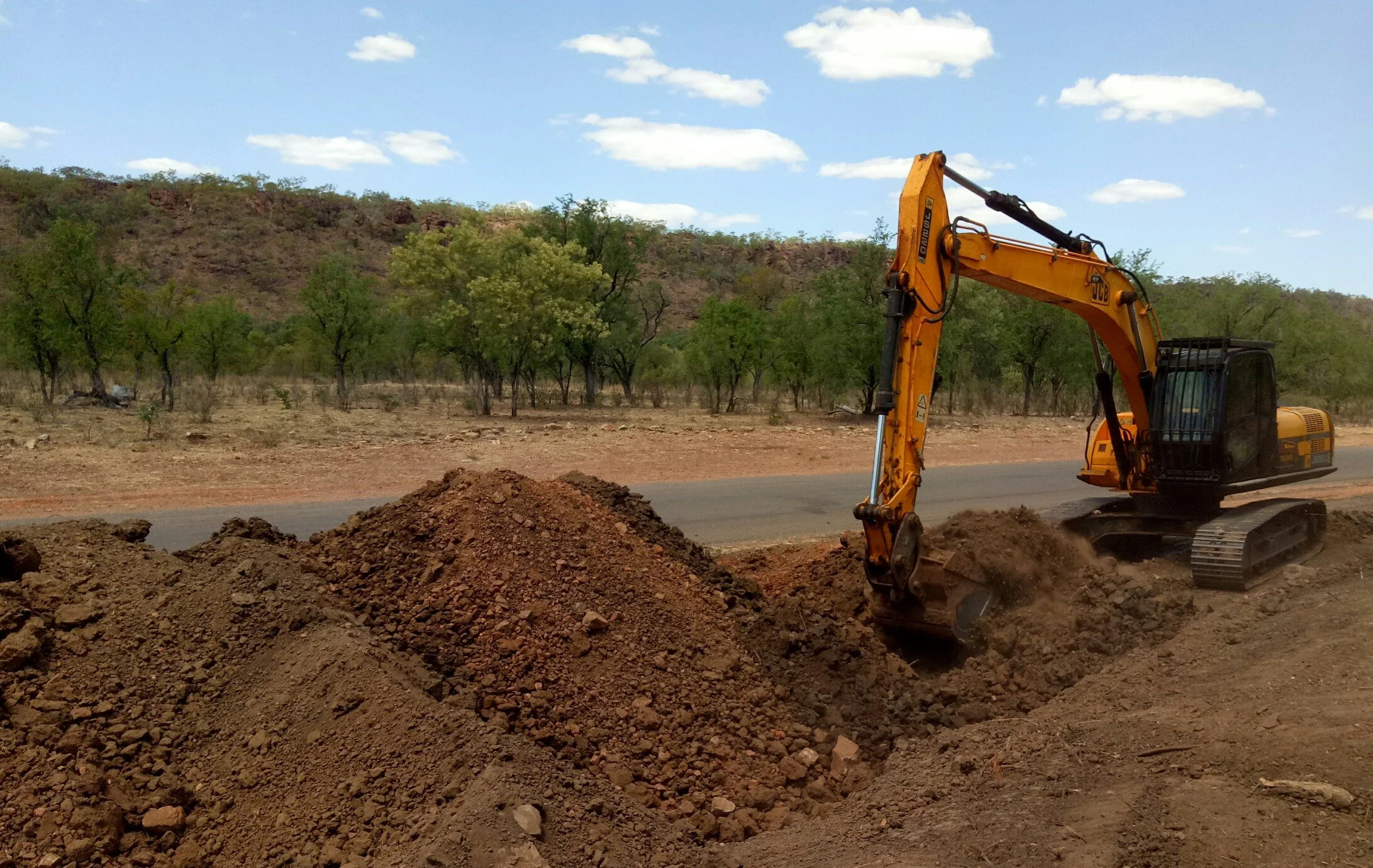 A construction excavator digging into dirt beside a rural road with trees and hills in the background and a partly cloudy sky.