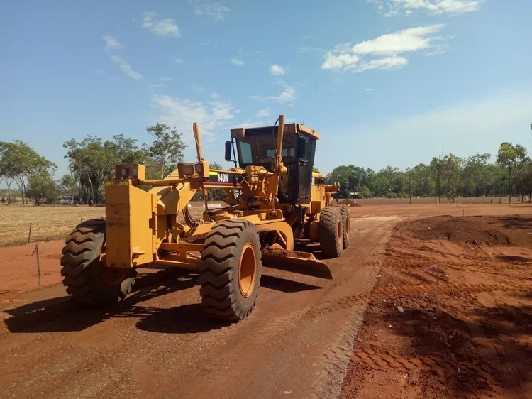 A yellow road grader working on a construction site with a dirt road and trees in the background.