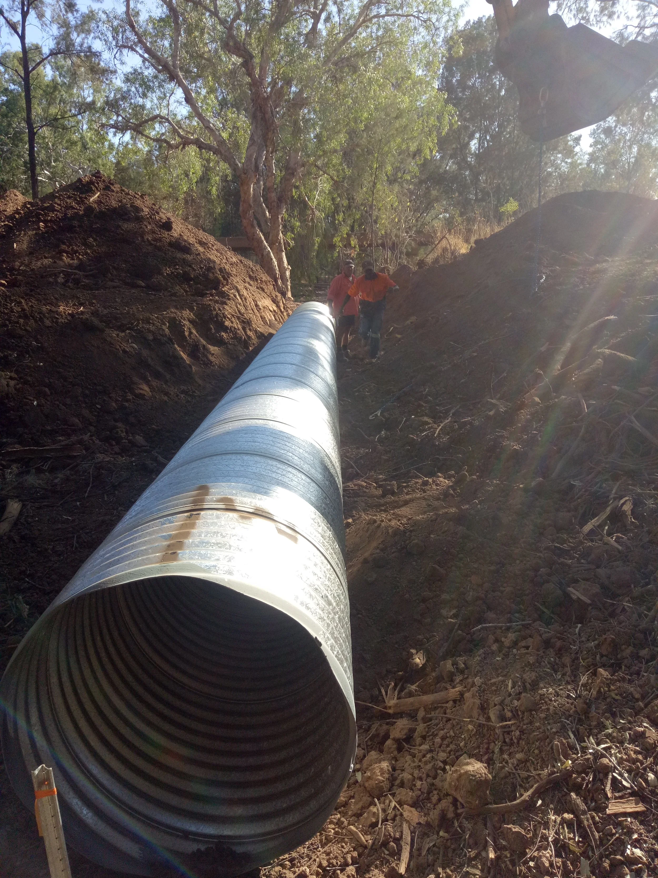 Workers installing a large black pipe in a trench surrounded by dirt and trees during daylight.