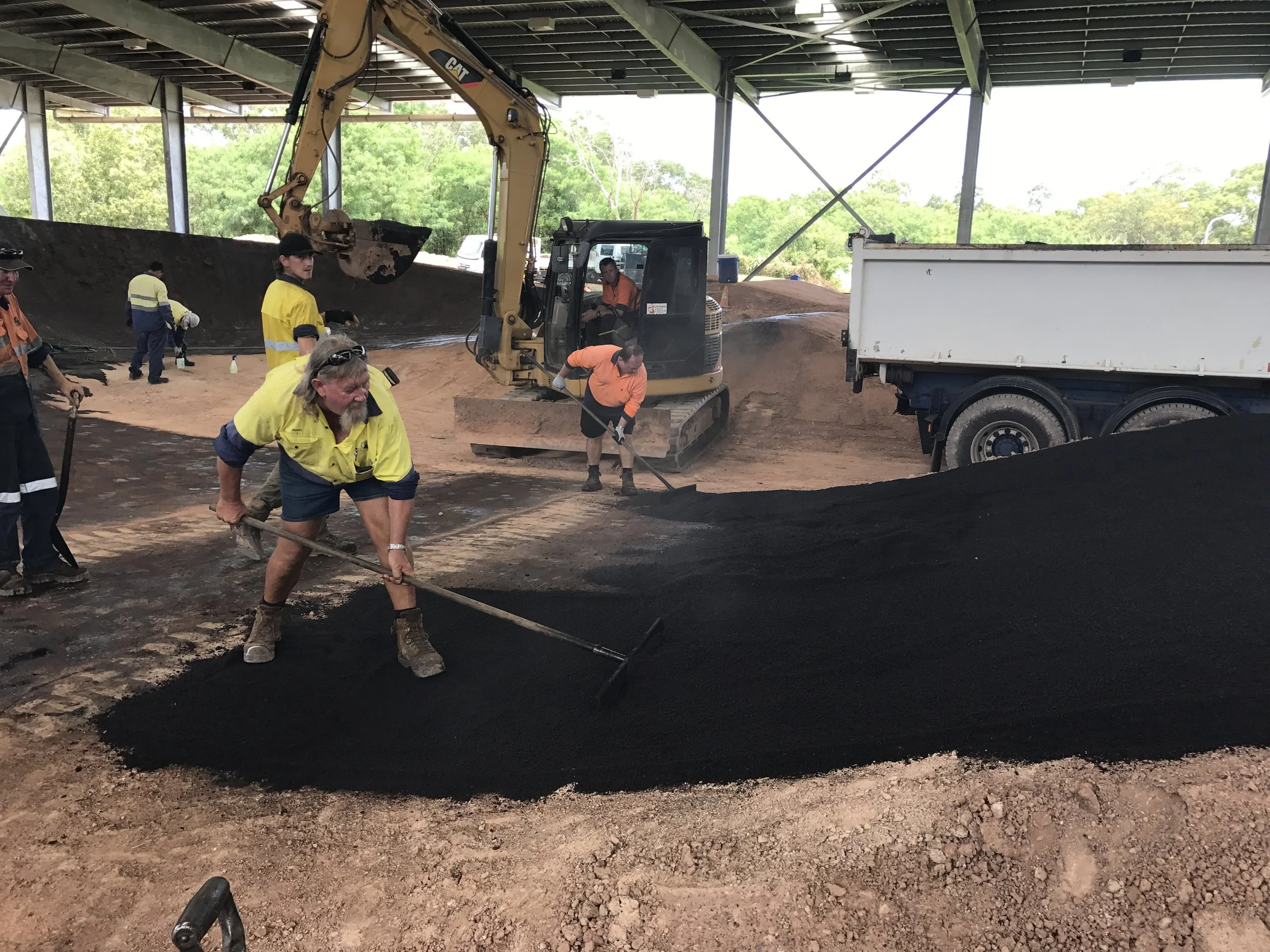 Construction workers spreading black asphalt inside a covered area, with road-building equipment and trucks in the background.