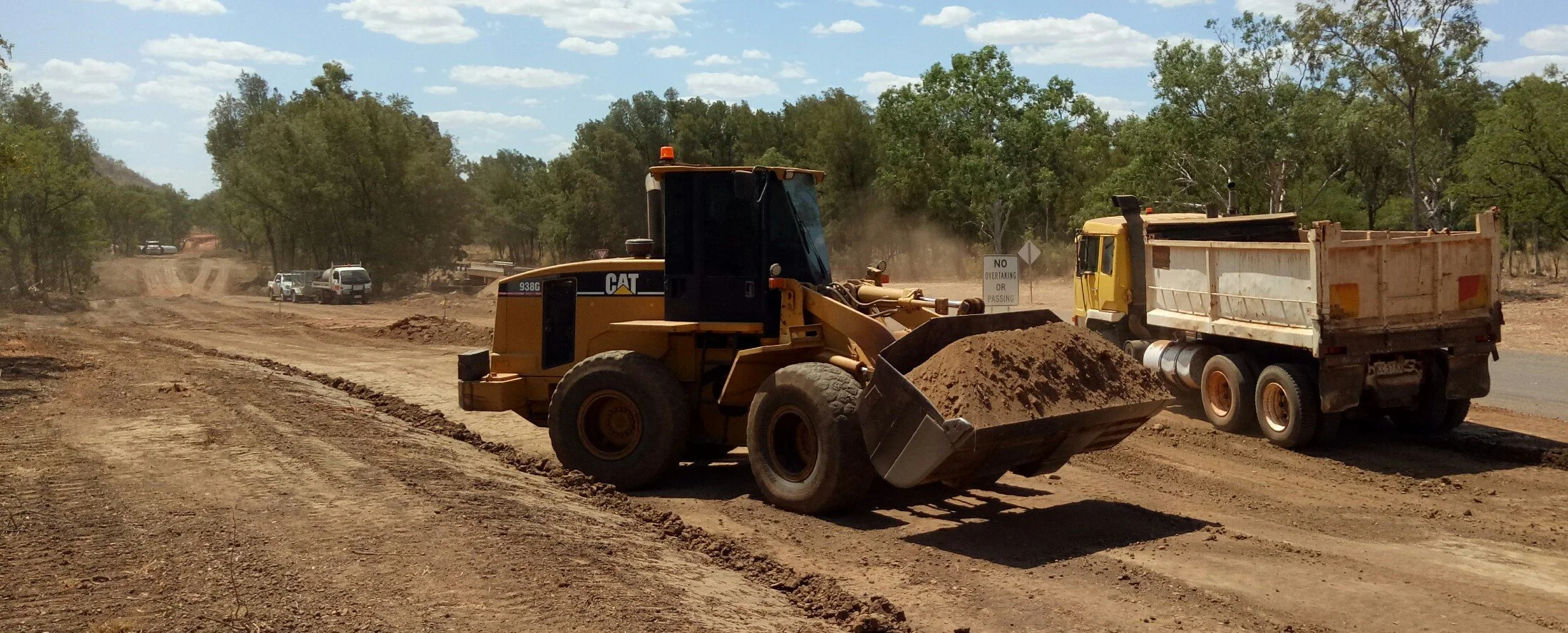 Construction site with a yellow CAT bulldozer pushing dirt and a yellow dump truck nearby, under a partly cloudy sky surrounded by trees.