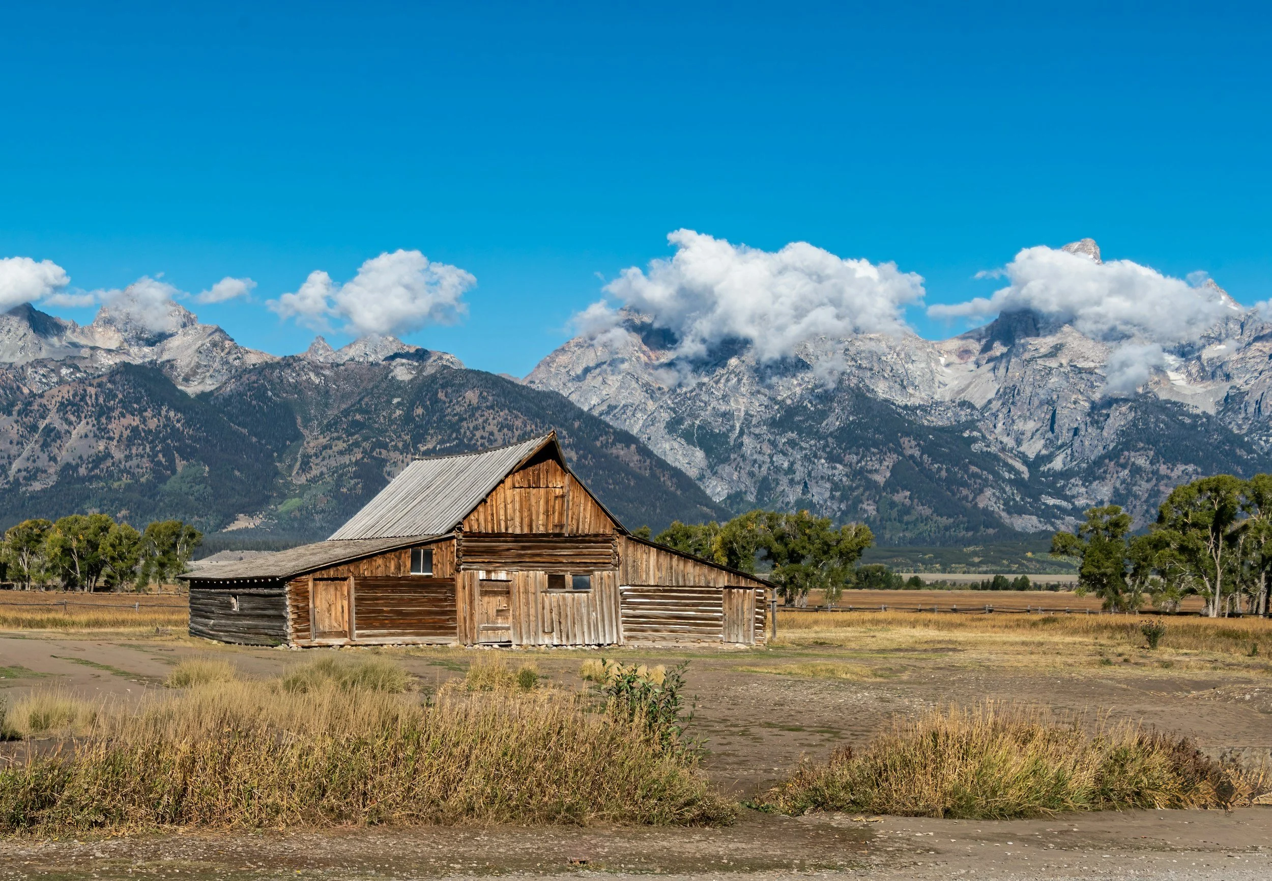 A rustic wooden barn in a vast field with mountains and clouds in the background under a bright blue sky.