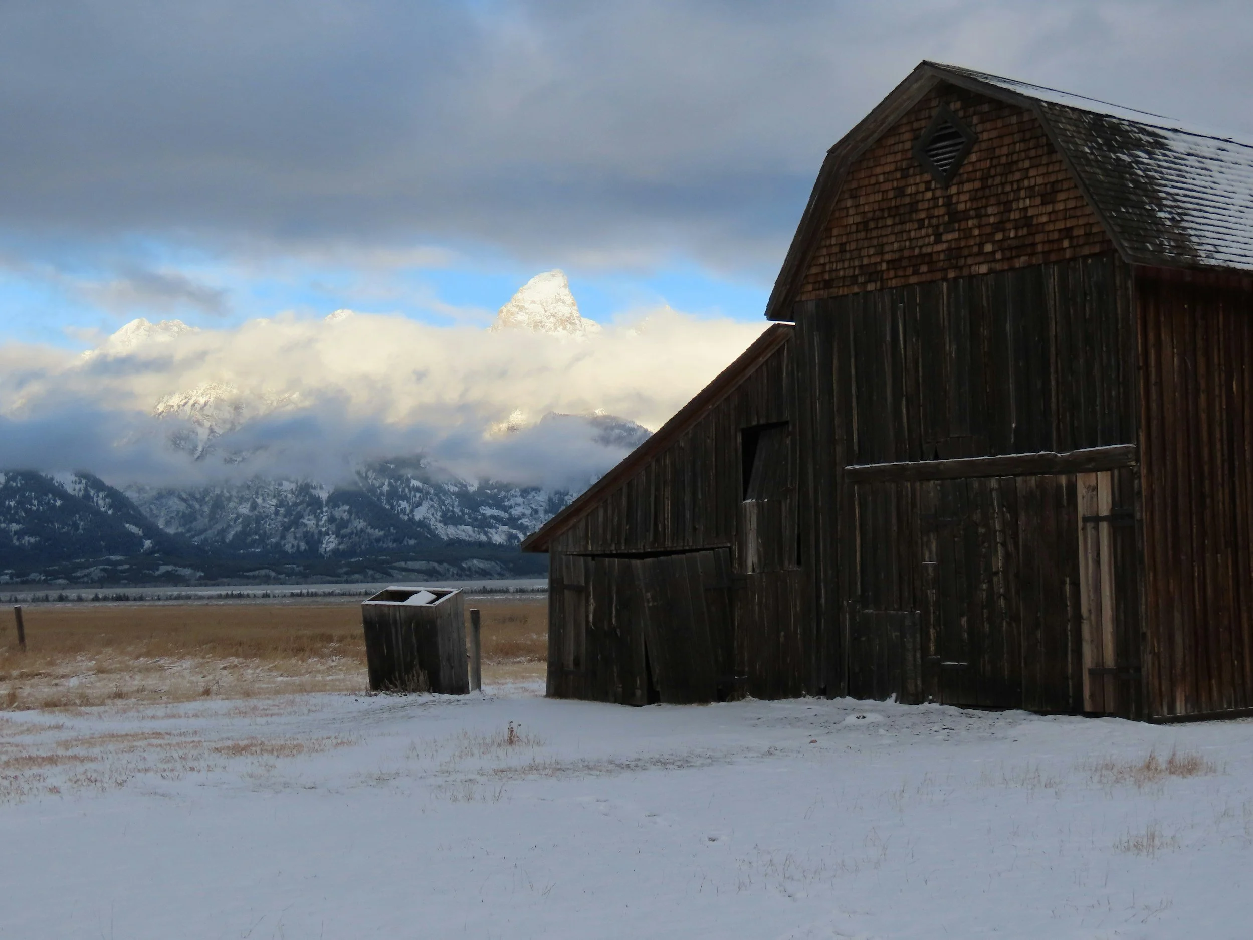 Old wooden barn in a snowy field with mountains in the background and clouds in the sky.