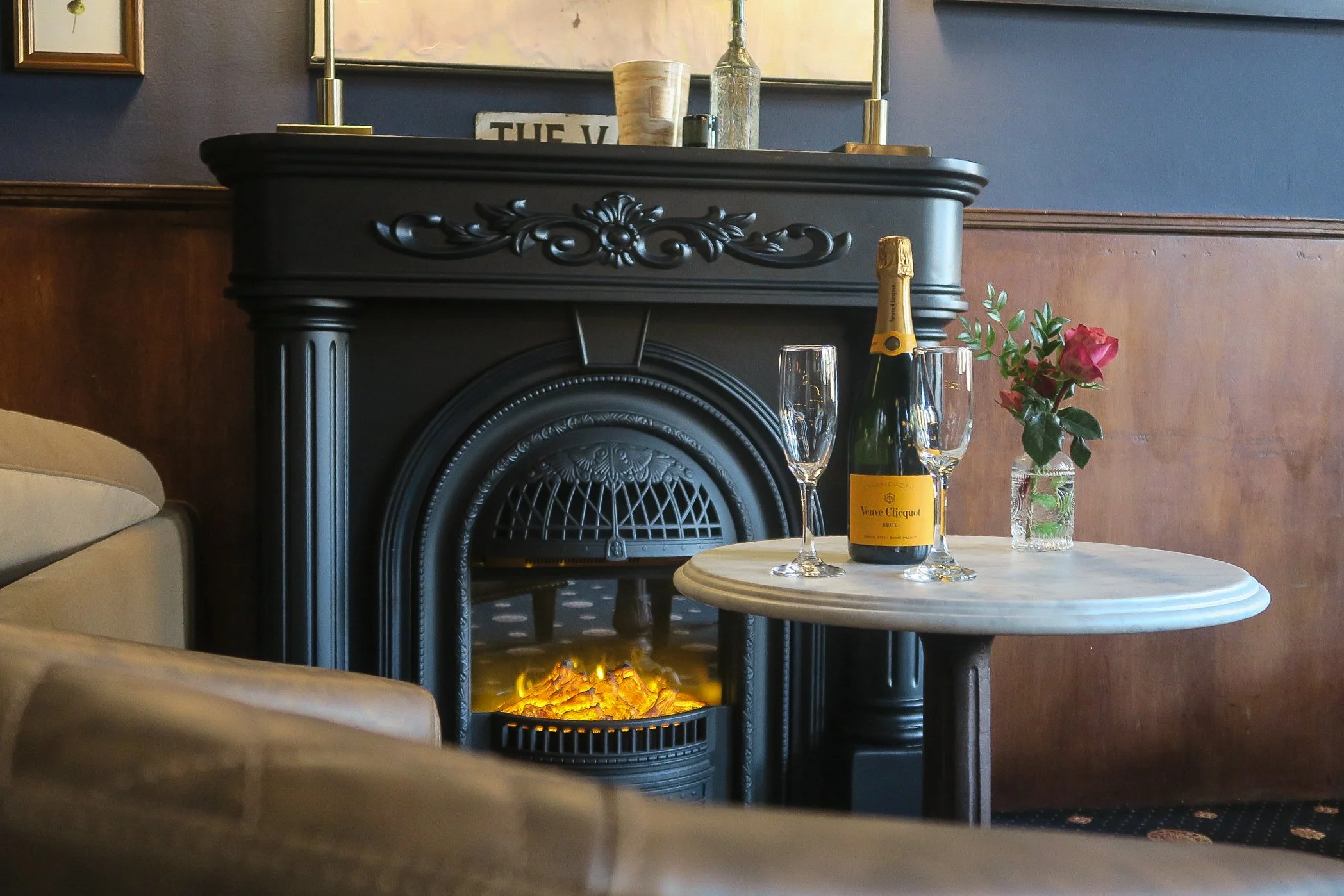 A cozy setting featuring a black ornate fireplace with a fire inside, a small round table with a bottle of Veuve Clicquot champagne, two champagne glasses, and a small vase with pink roses.