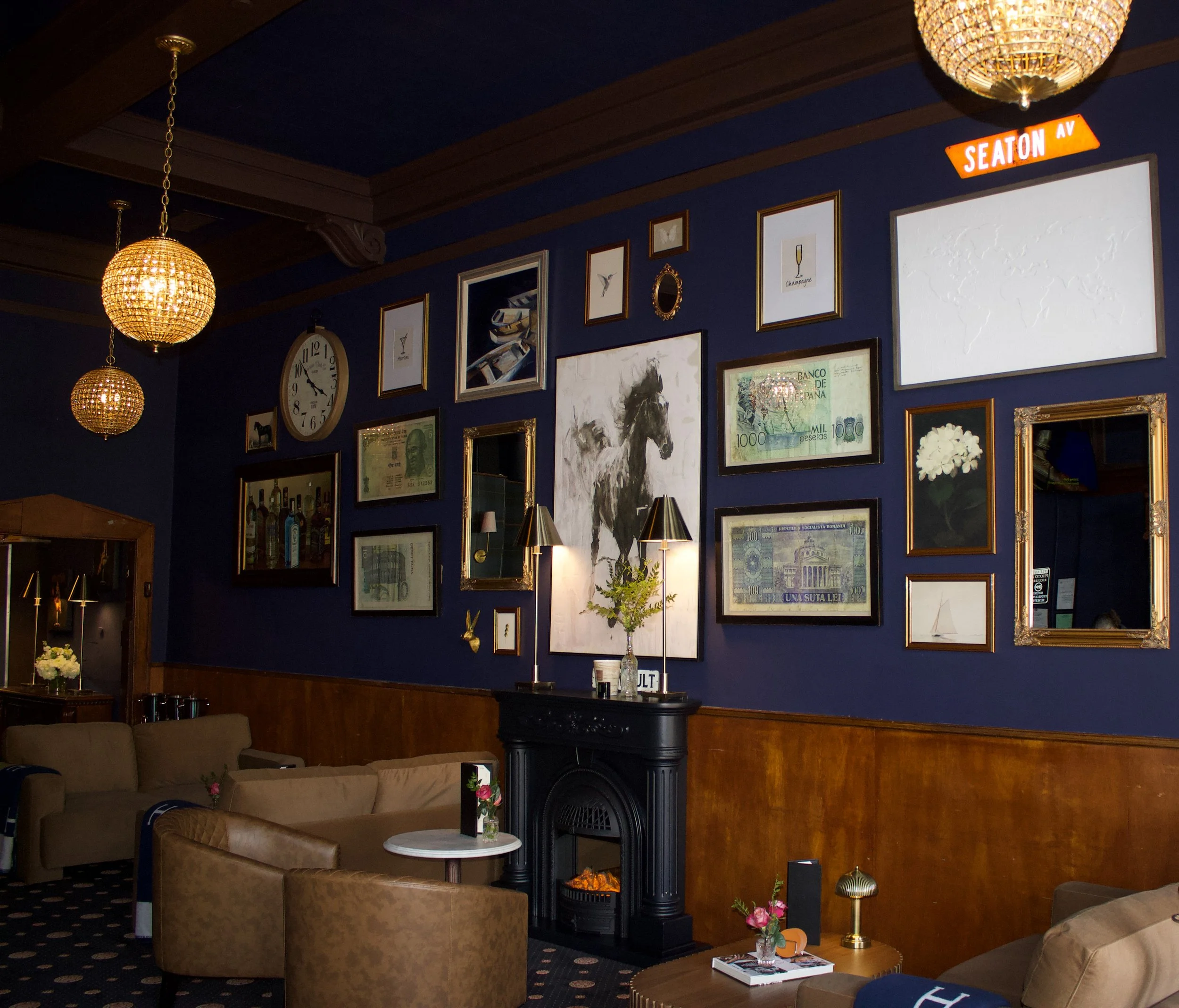 Interior of a cozy lounge with dark blue walls decorated with framed pictures and artwork, including currency notes and paintings. There is a fireplace with a small plant on top, surrounded by beige sofas and small tables with flowers. Two hanging spherical lights and a wall clock are also visible.