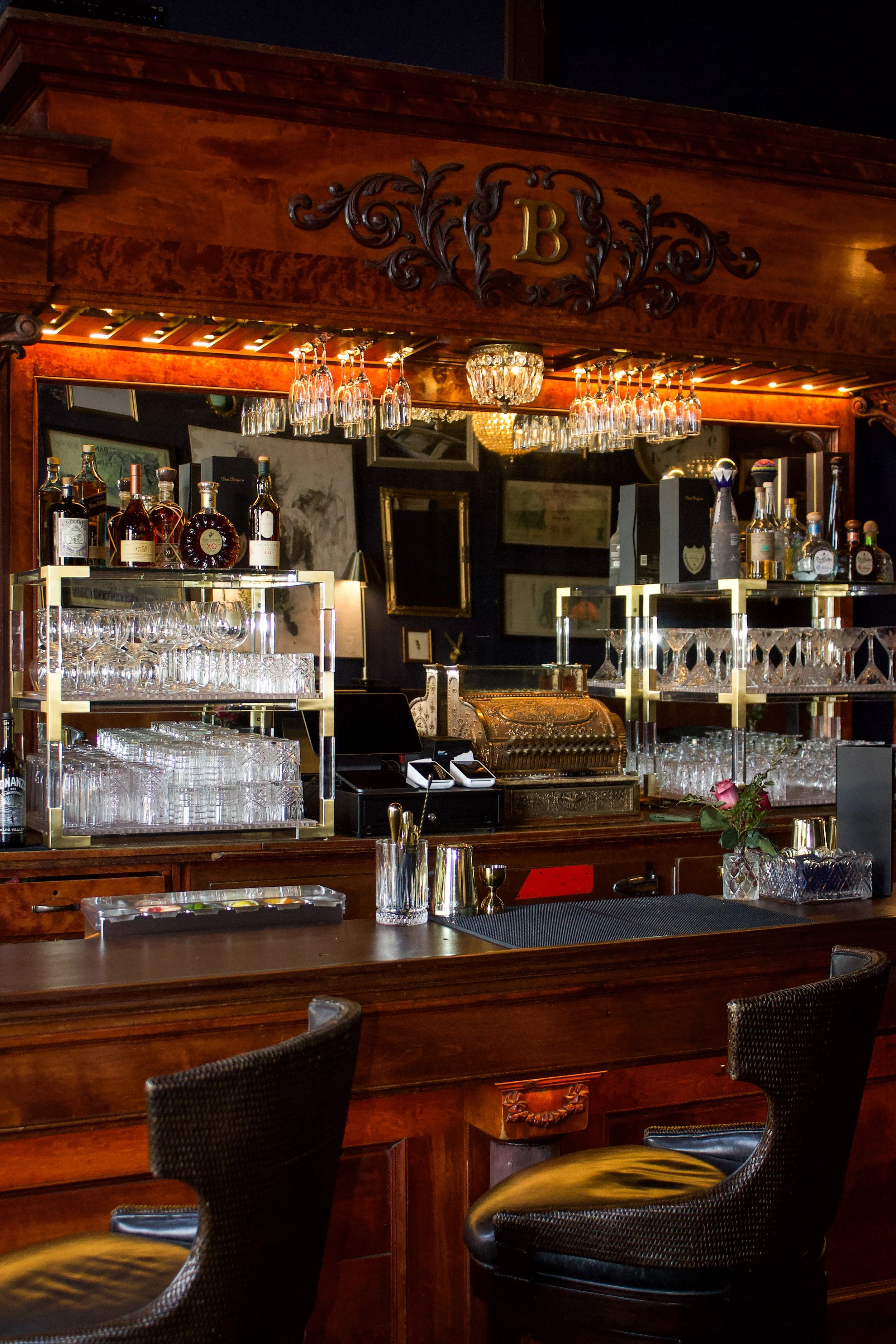 A well-lit bar with wooden furnishings, glassware racks, bottles of liquor, and a rose in a vase on the counter.