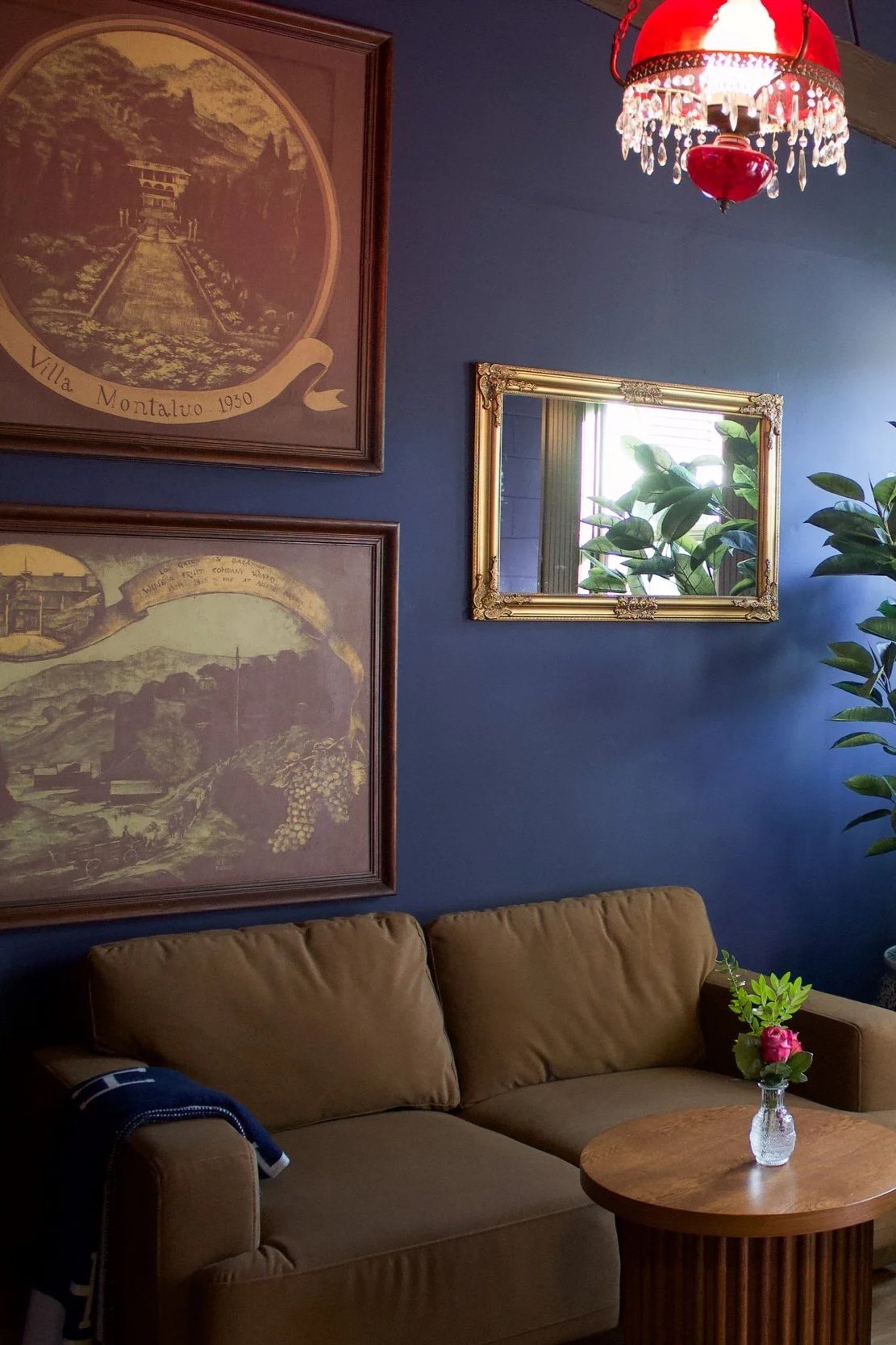 Cozy living room with a brown sofa, a round wooden coffee table with a glass vase holding pink flowers and green leaves. A mirror with a gold frame hangs on a blue wall, reflecting a window with blinds. Two framed signs or pictures with scenic illustrations and text are above the sofa. A red and white vintage-style chandelier hangs from the ceiling, and a large green plant is near the window.