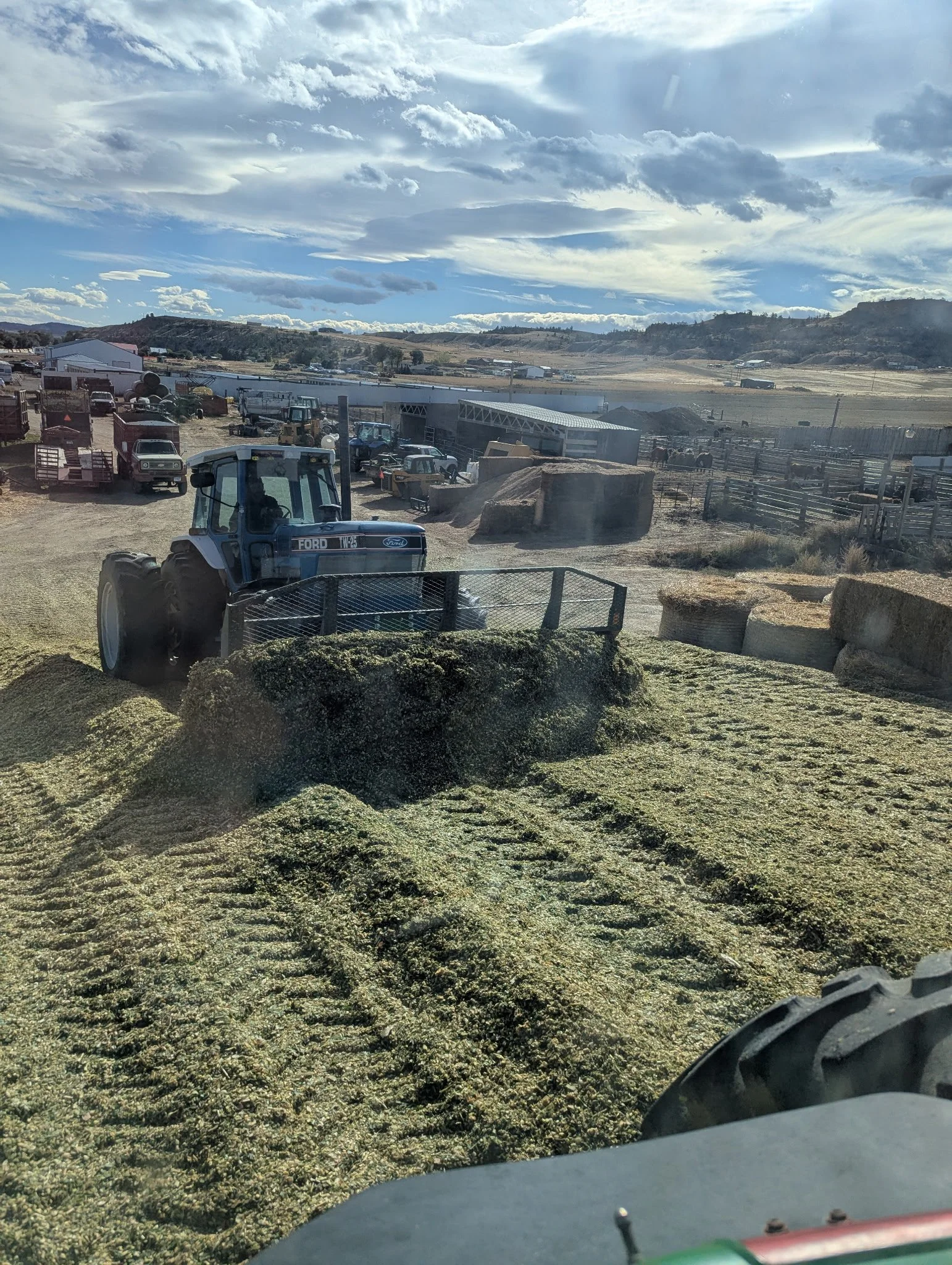Farming equipment and hay bales in a rural landscape, with a tractor in the foreground and a cloudy sky above.