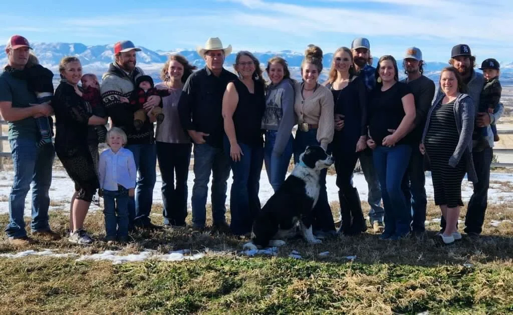 Group of people standing outdoors on a snowy hill with mountains in the background, posing with a large black and white dog.
