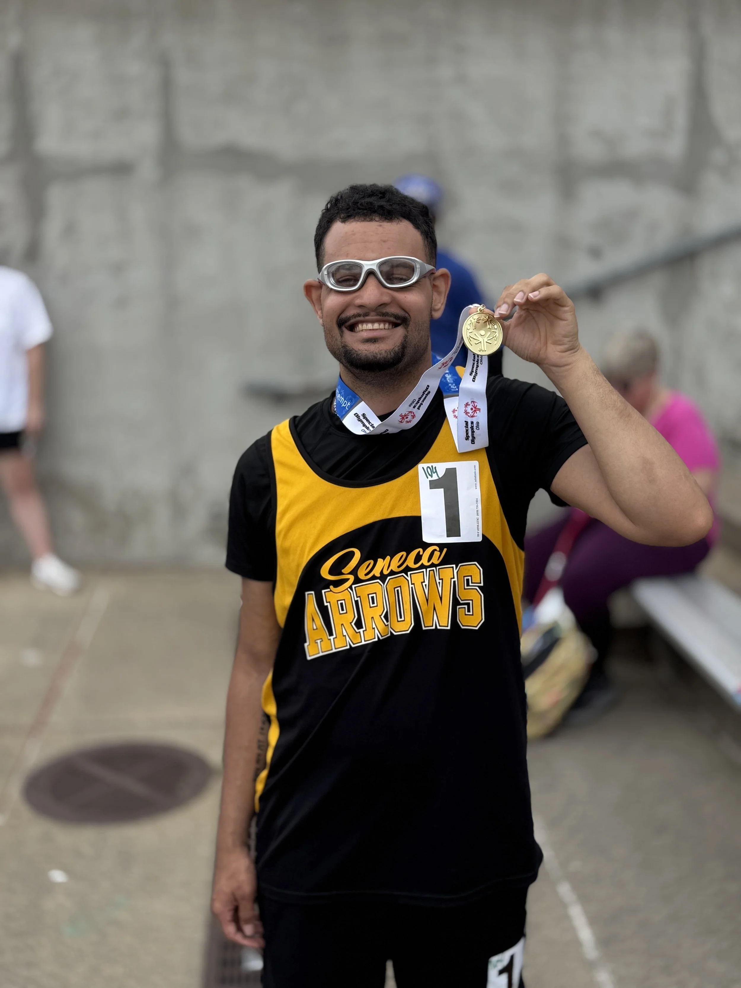 A man smiling and holding up a gold medal at a race event, wearing goggles, a black and yellow jersey with 'Seneca Arrows' written on it, and a race bib with the number 1.
