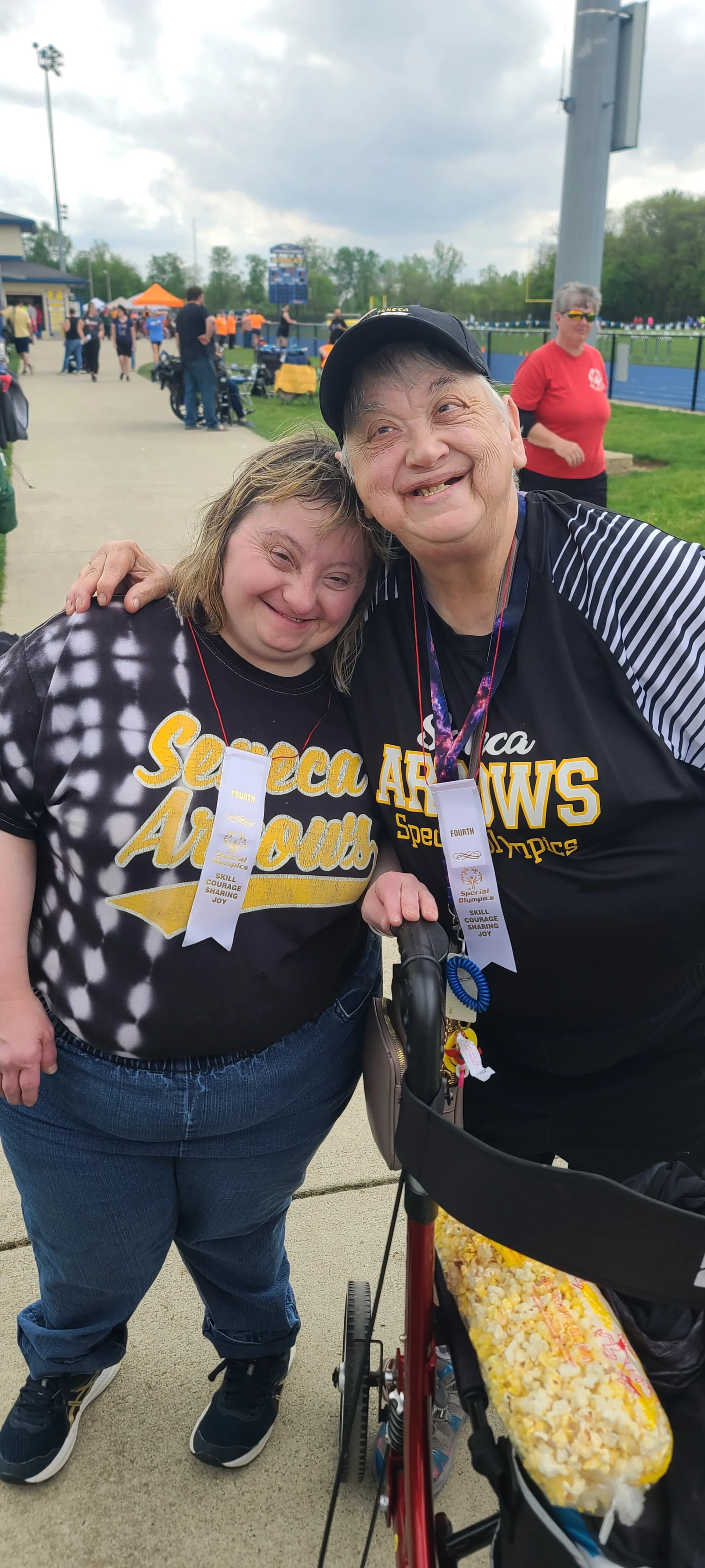 Two women wearing matching 'Special Olympics' shirts with medals, smiling and hugging at an outdoor event, with a track and people in the background.