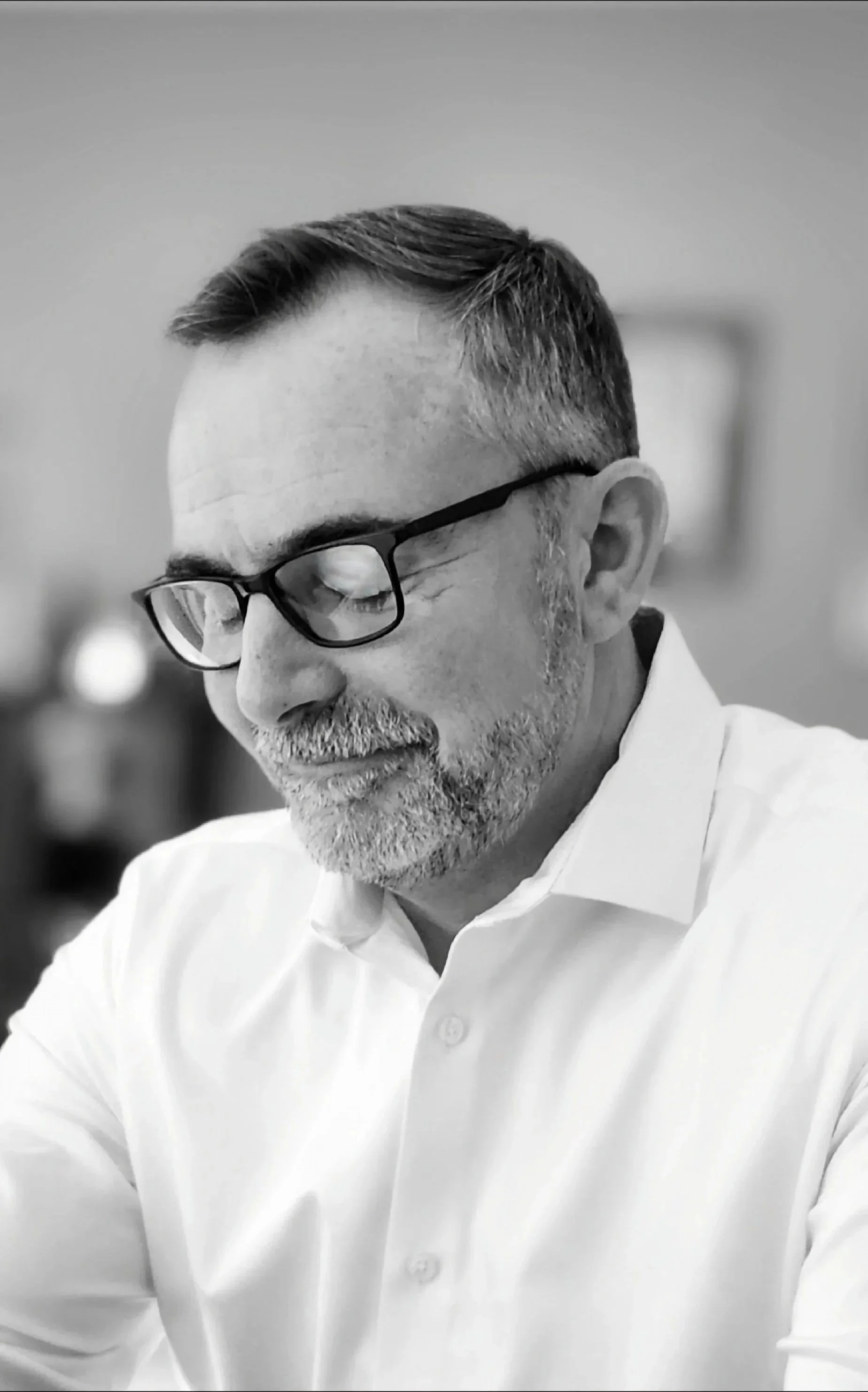 Black and white portrait of Sean Riggin wearing glasses and a white shirt, looking down thoughtfully.