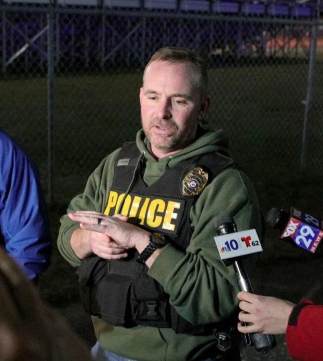 Portrait of Sean Riggin, while Chief of Police, wearing a vest with 'POLICE' in yellow letters, being interviewed by reporters outside at night.