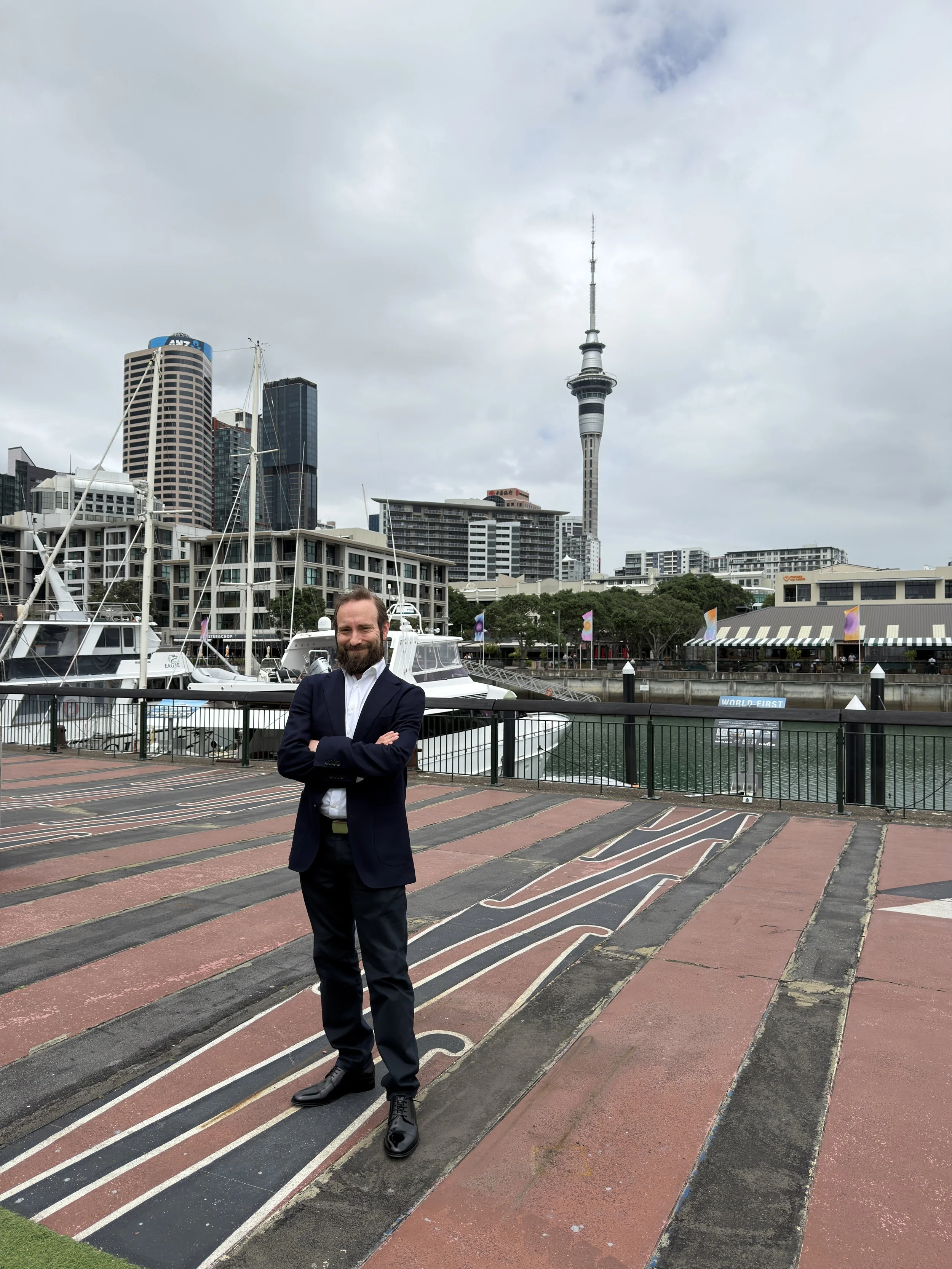 A man in a suit standing with arms crossed near a marina with yachts and city buildings, including a tall tower in the background, under a cloudy sky.