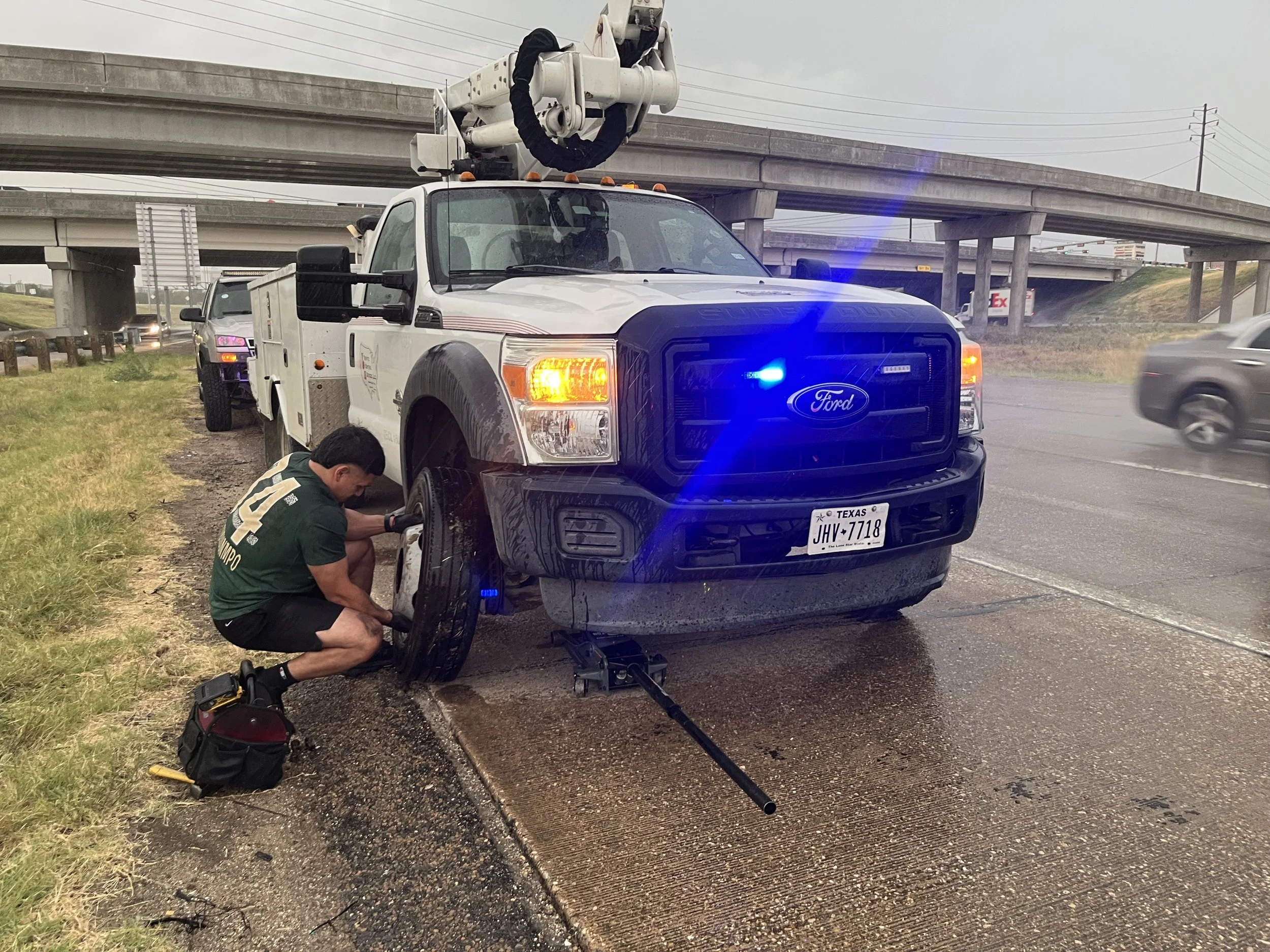 A mobile tire service emergency worker changing a flat tire on a white Ford pickup truck on the side of a wet highway, with a highway overpass overhead and other vehicles passing by.