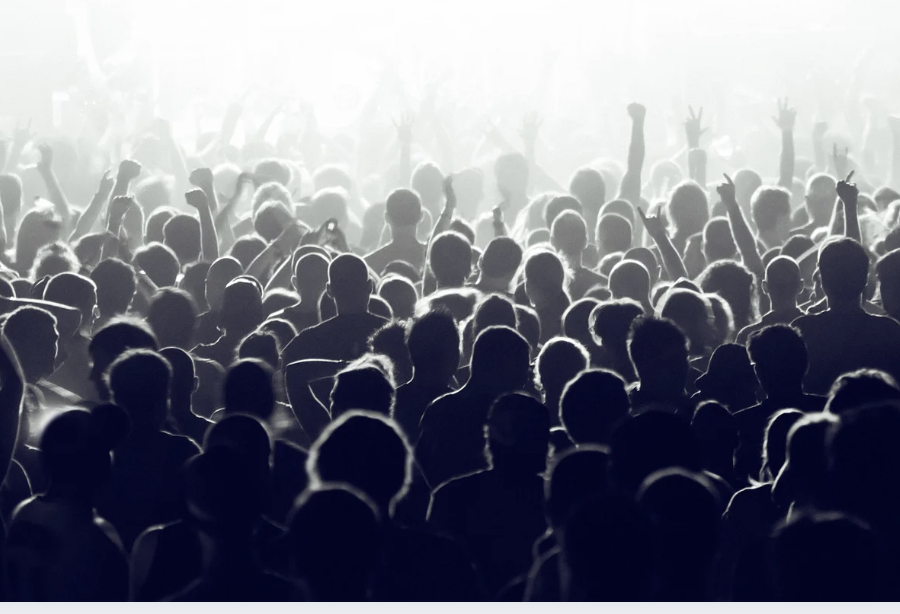 Crowd of people at a concert or event with stage lighting in the background.